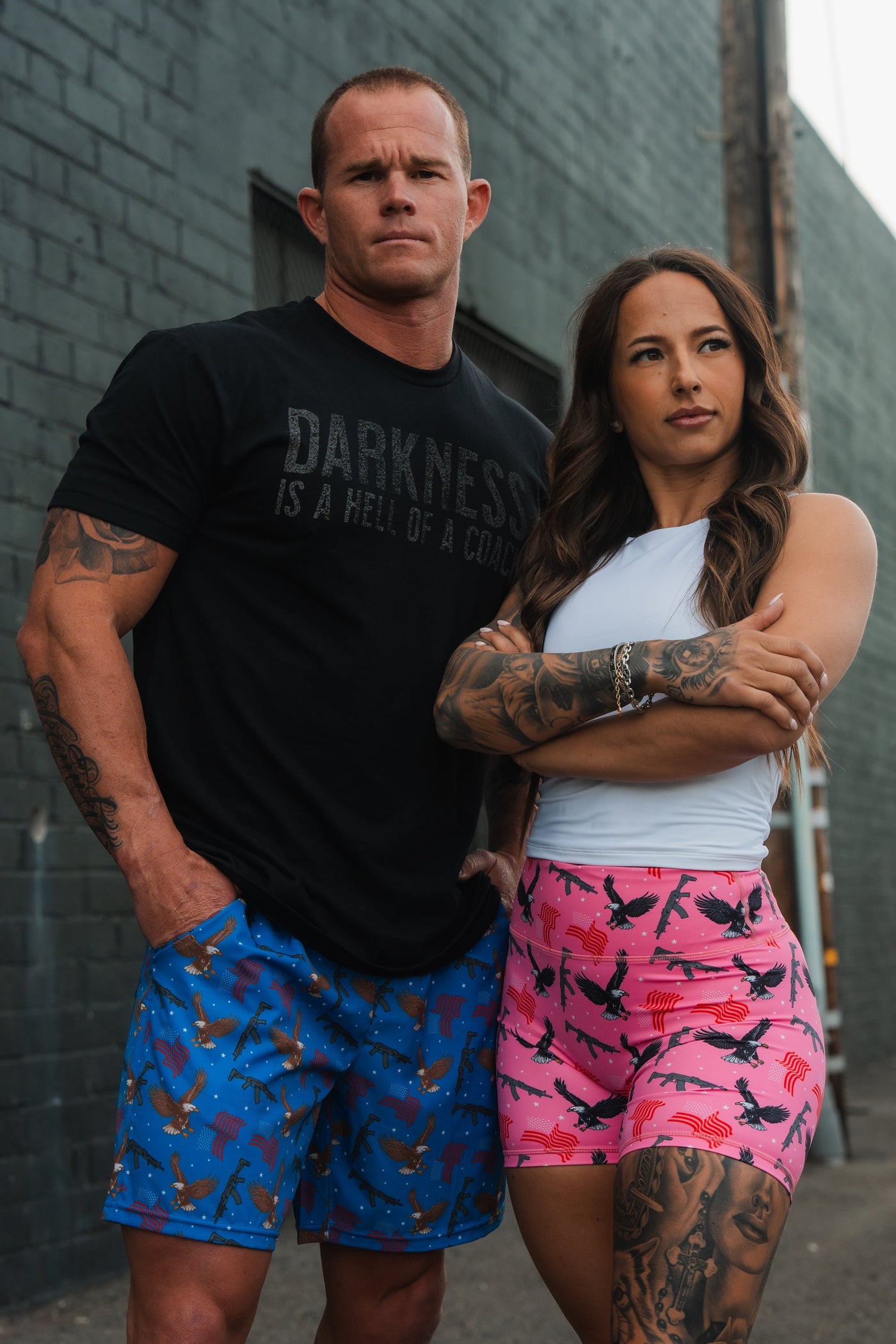 A man in a black shirt and blue shorts stands next to a woman in a white top and pink shorts. Both have tattoos and serious expressions, posing in an urban alley.