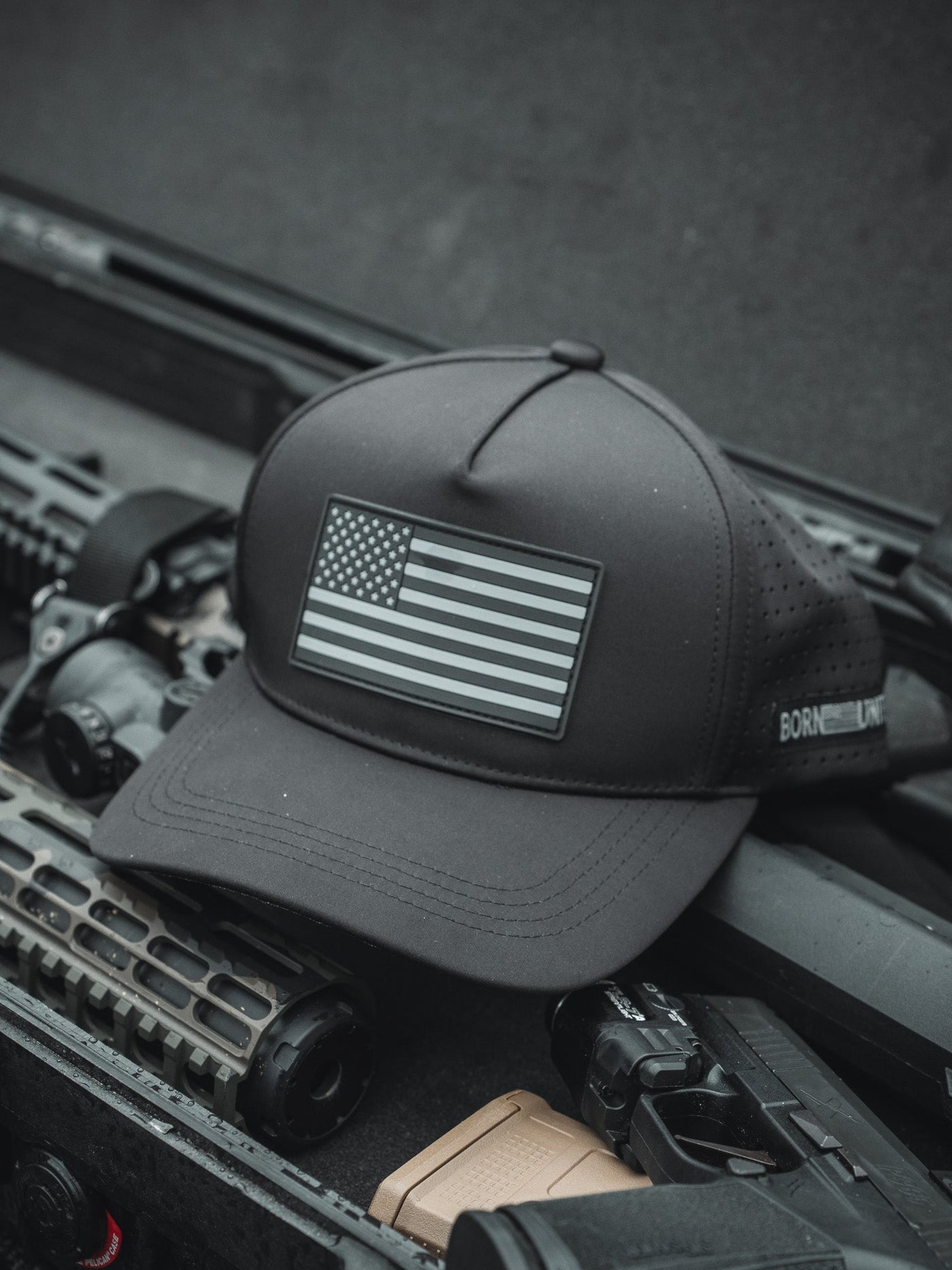 A black baseball cap with a gray American flag patch rests on top of tactical gear, including a rifle and firearm accessories.