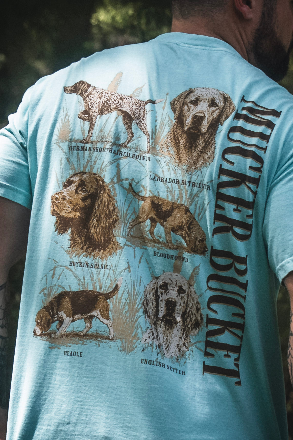 A man wearing the Dog DNA Premium Tee by Mucker Bucket, a premium shirt showcasing illustrations and names of six dog breeds—German Shorthaired Pointer, Labrador Retriever, Cocker Spaniel, Bloodhound, Beagle, and English Setter.