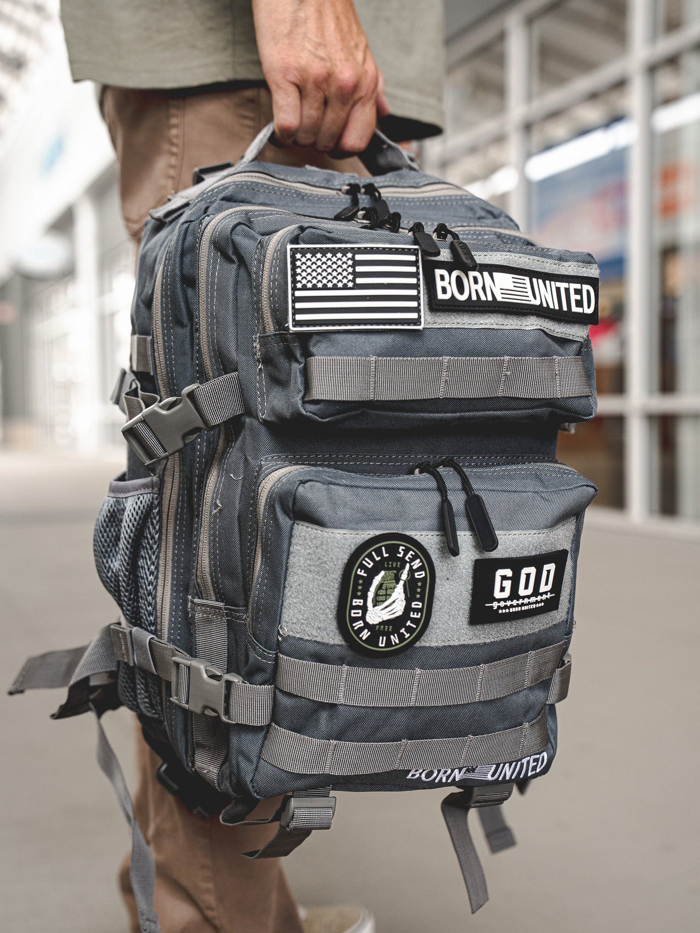 A person holds a gray tactical backpack adorned with patches, including an American flag, BORN UNITED, and GOD FULL SEND UNITED. The background shows a blurred outdoor walkway.