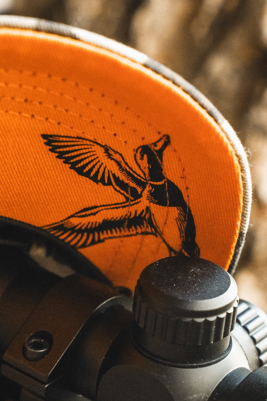 Close-up of a hat with an orange underside featuring a black illustration of a flying duck. A portion of what appears to be a camera or optical device is visible in the foreground.