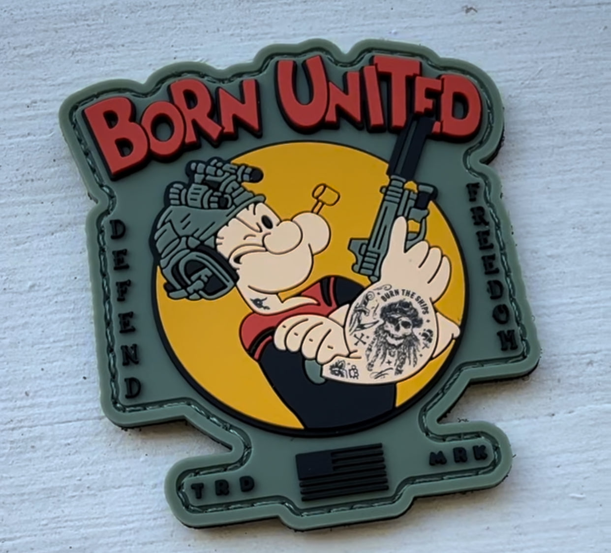 A patch featuring Popeye wearing military gear and holding a rifle, with “BORN UNITED” in red above him and “DEFEND FREEDOM” on the sides. Popeye has a skull tattoo and a yellow background circle.