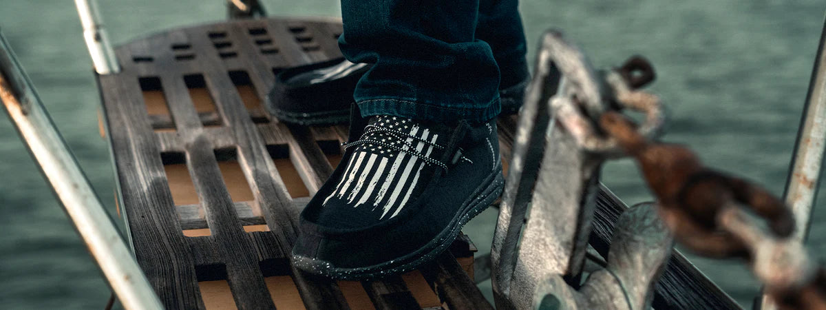 Close-up of a person standing on a wooden surface wearing black shoes with white vertical stripes and polka dot laces, with water and metal boat parts in the background.