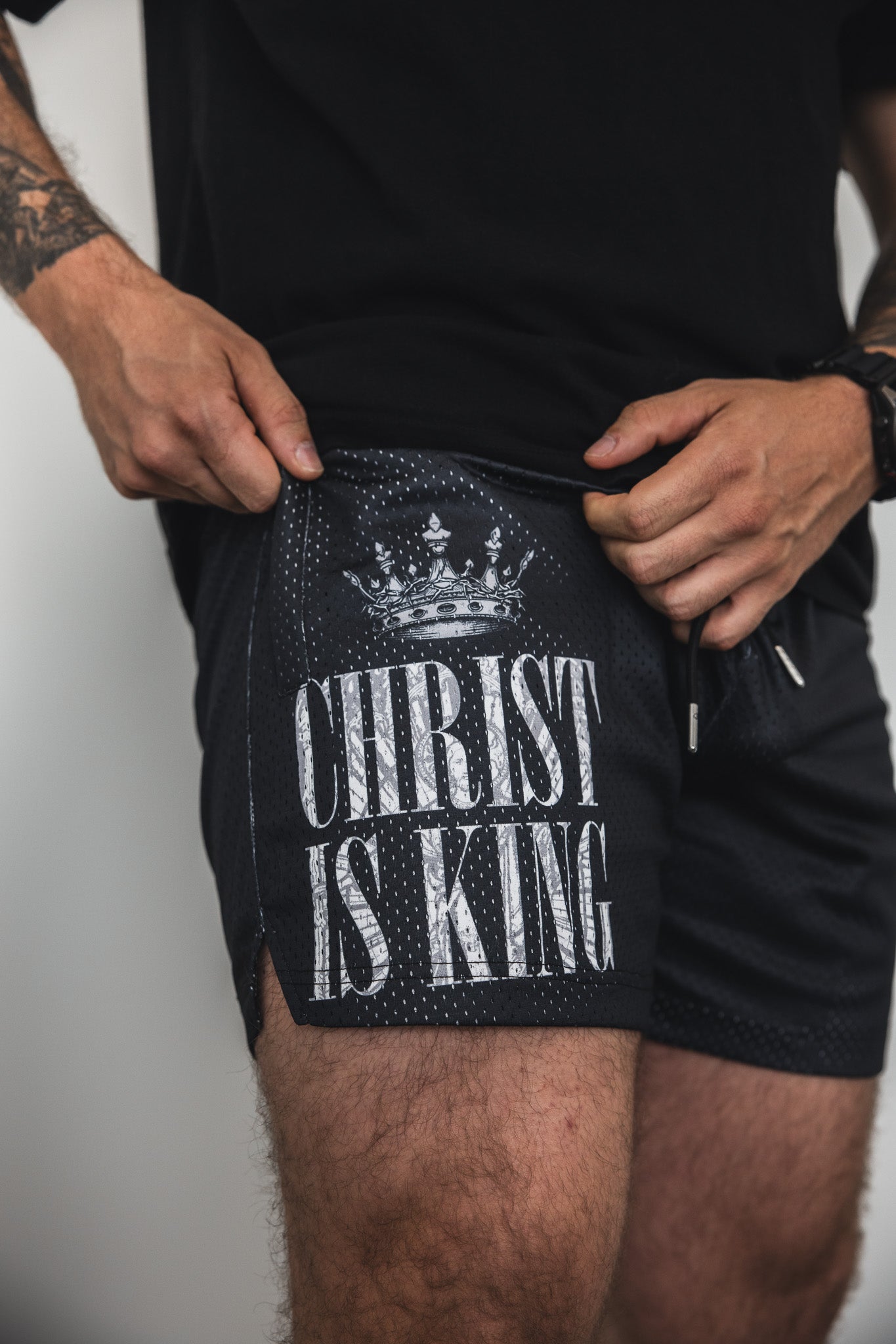 A person wearing black shorts with a crown graphic and the text CHRIST IS KING printed on the side, holding the waistband with both hands.