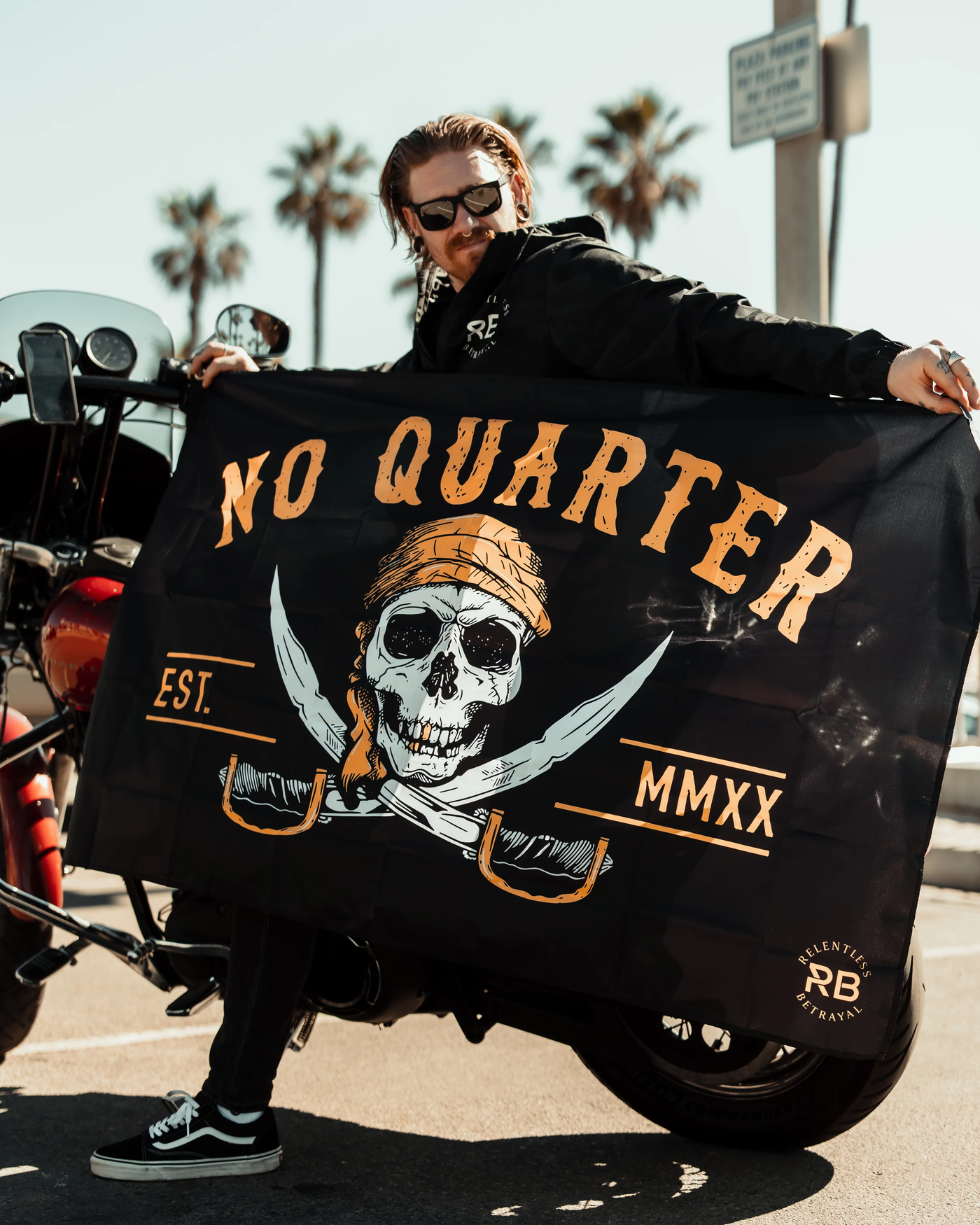 A man in sunglasses and a hoodie holds a flag that reads NO QUARTER with a skull in a bandana and crossed swords. A motorcycle and palm trees are visible in the background.