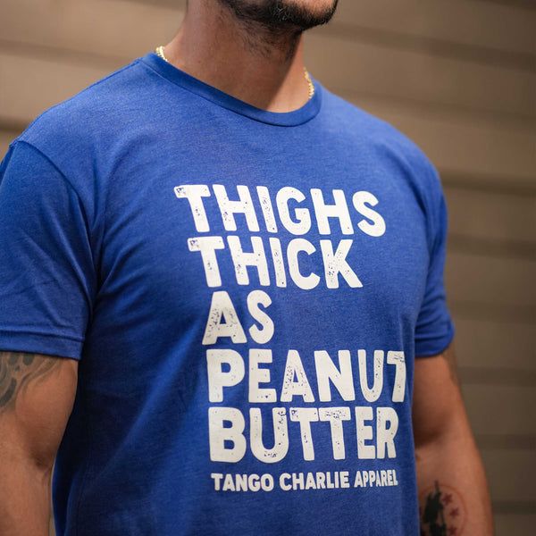 A person wearing a blue T-shirt with white text that reads, THIGHS THICK AS PEANUT BUTTER TANGO CHARLIE APPAREL. The persons face is not visible.
