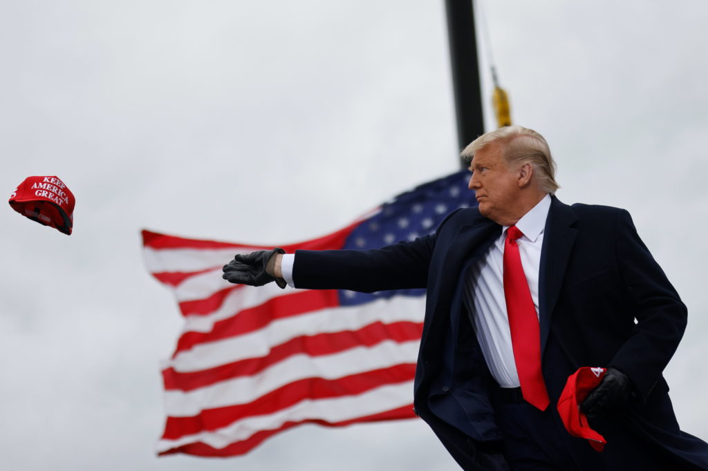 A man in a dark coat and red tie throws a red hat while holding another, with a large American flag waving in the background against a cloudy sky.