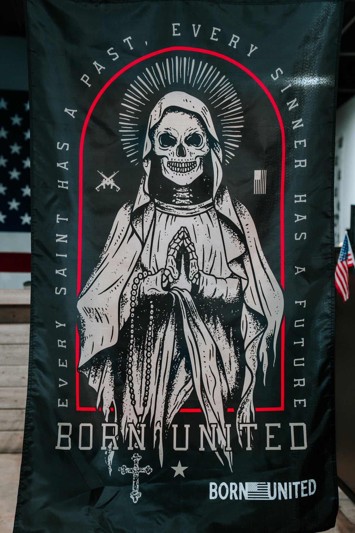 The Born United Private Label Saints and Sinners Flag is a black polyester flag featuring a skeletal saint in a robe with halo and praying hands, with the phrases EVERY SAINT HAS A PAST, EVERY SINNER HAS A FUTURE and BORN UNITED. An American flag appears in back.