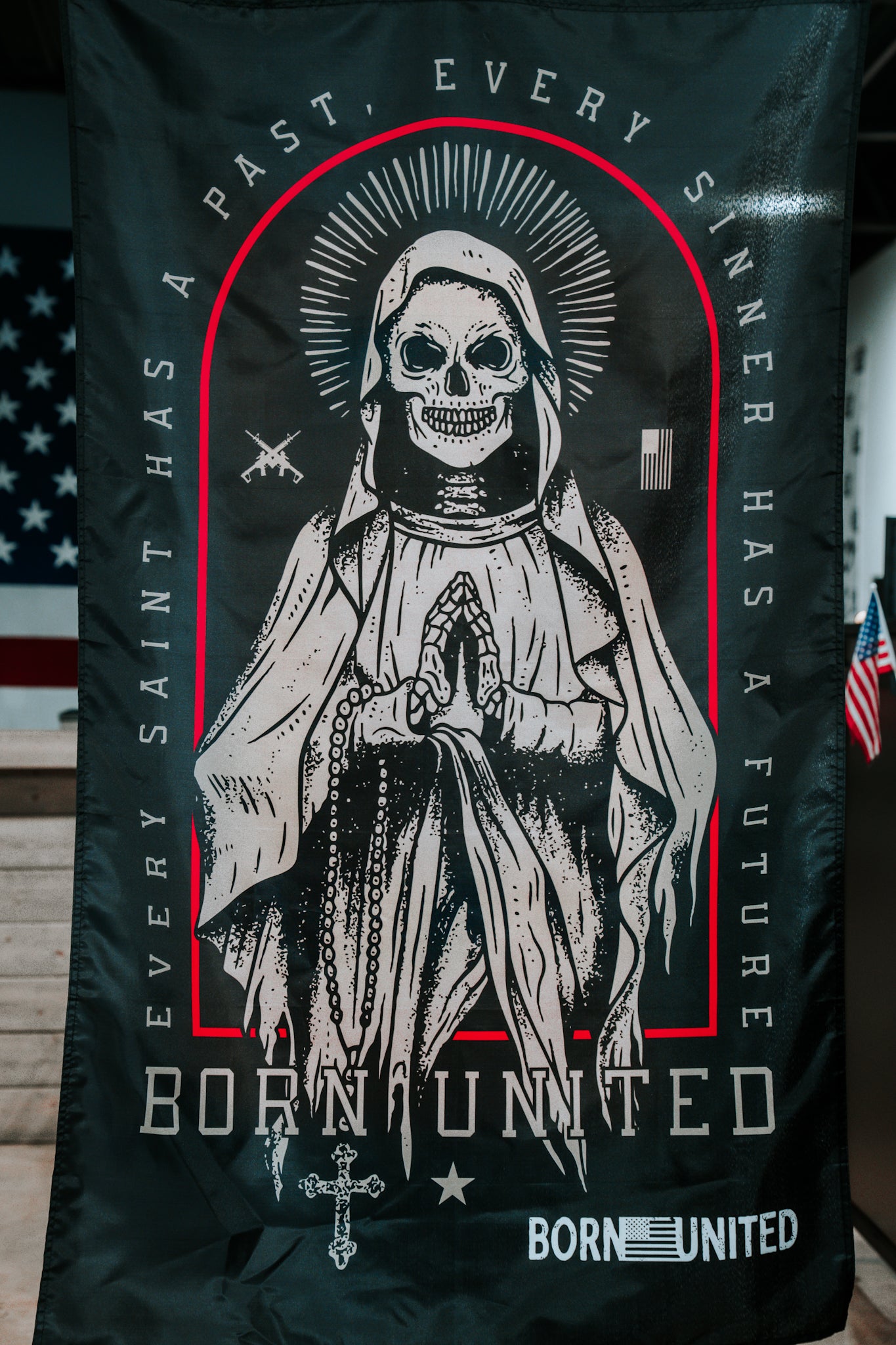 The Born United Private Label Saints and Sinners Flag is a black polyester flag featuring a skeletal saint in a robe with halo and praying hands, with the phrases EVERY SAINT HAS A PAST, EVERY SINNER HAS A FUTURE and BORN UNITED. An American flag appears in back.
