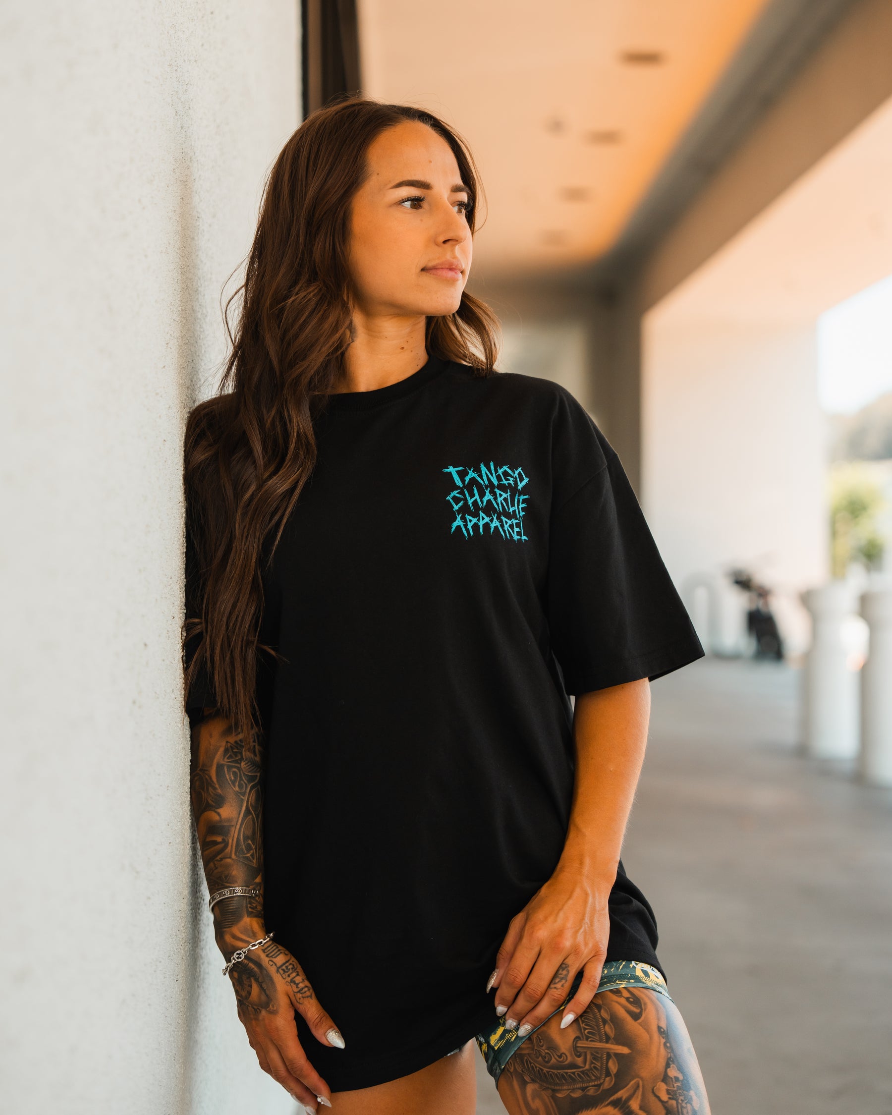 A woman with long brown hair and arm tattoos leans against a white wall in the sun, wearing the Tango Charlie Apparel Psych Ward Premium Oversized Tee—black cotton with Tango Charlie Apparel in blue lettering. She gazes to the side.
