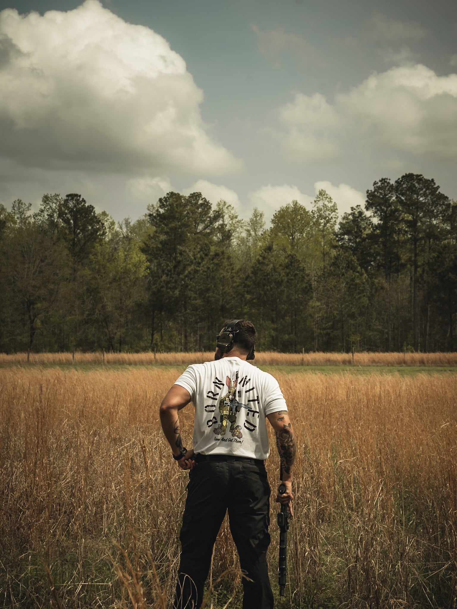 In a field of tall grass under a cloudy sky, a person stands facing away, holding a stick. They wear Born United Private Labels Battle Bunny white shirt featuring a unique design on the back, while tall trees line the horizon like guests at nature’s Easter dinner.
