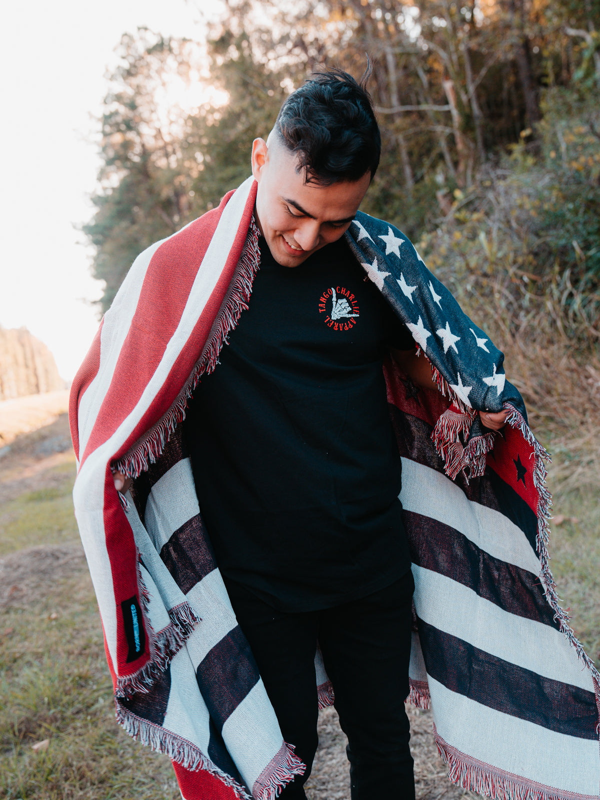 The person stands outdoors, wrapped in the Born United Private Labels American Flag Jacquard Blanket, smiling as they look down. They are dressed in a black t-shirt and pants, with trees and a clear sky in the background.
