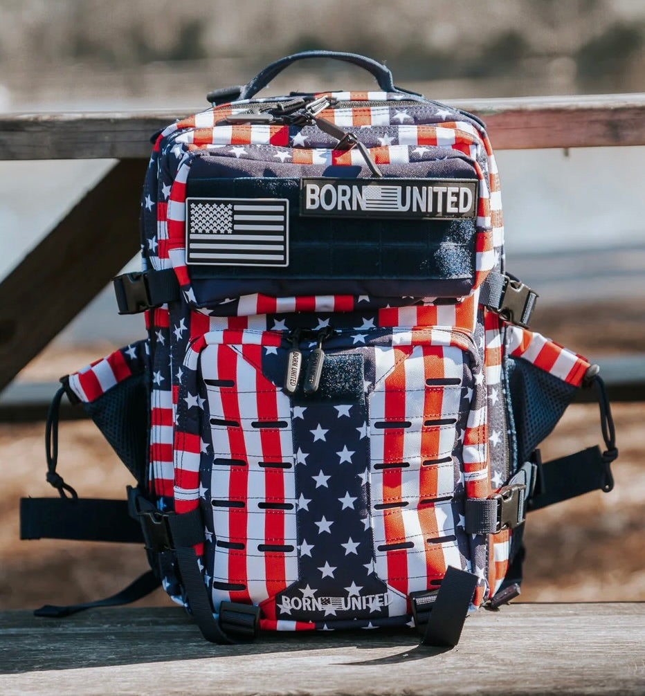 The Premium Tac-Pac Patch Bundle by Born United Private Label is a backpack with an American flag pattern, stars, red, white and blue stripes, plus BORN UNITED and American flag patches displayed on a wooden outdoor bench.