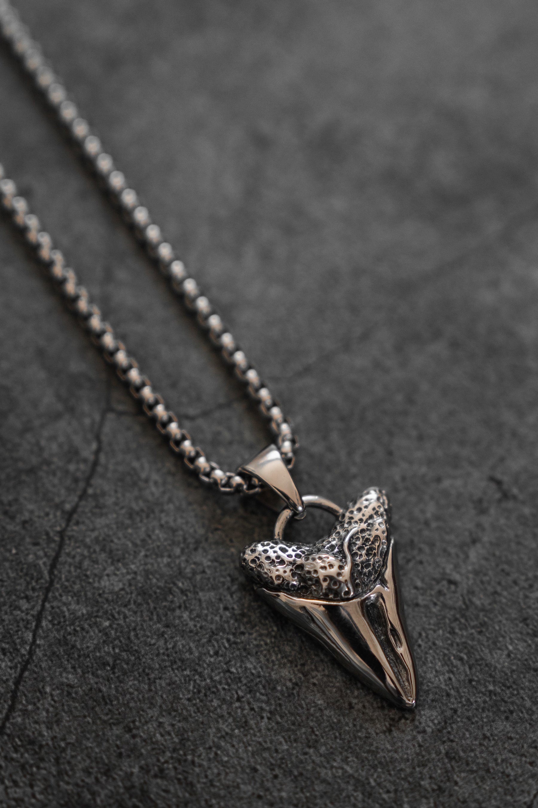 The Relentless Predator Necklace by Relentless Betrayal, crafted from stainless steel, lies on a textured dark surface. Its metallic pendant mimics a fossilized shark tooth with detailed etchings, embodying the relentless ocean predators essence.