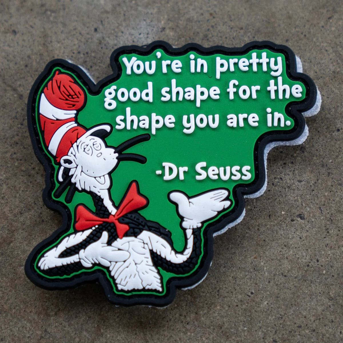 The St. Seuss PVC Patch by Tango Charlie Apparel, featuring the Cat in the Hat and the quote “You’re in pretty good shape for the shape you are in,” is shown on a green background atop a gray surface.