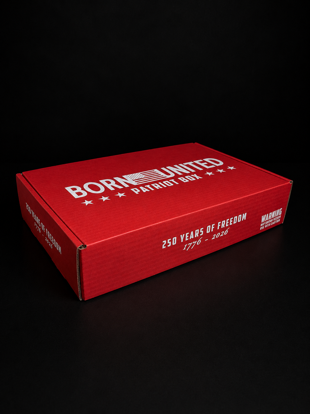 The Born United Patriot Box by Born United Private Label is a red cardboard box with “250 Years of Freedom 1776-2026” and “Warning: Contents May Cause Patriotism” on the front, marking America's 250th Birthday. Limited Edition Tees inside. Black background.