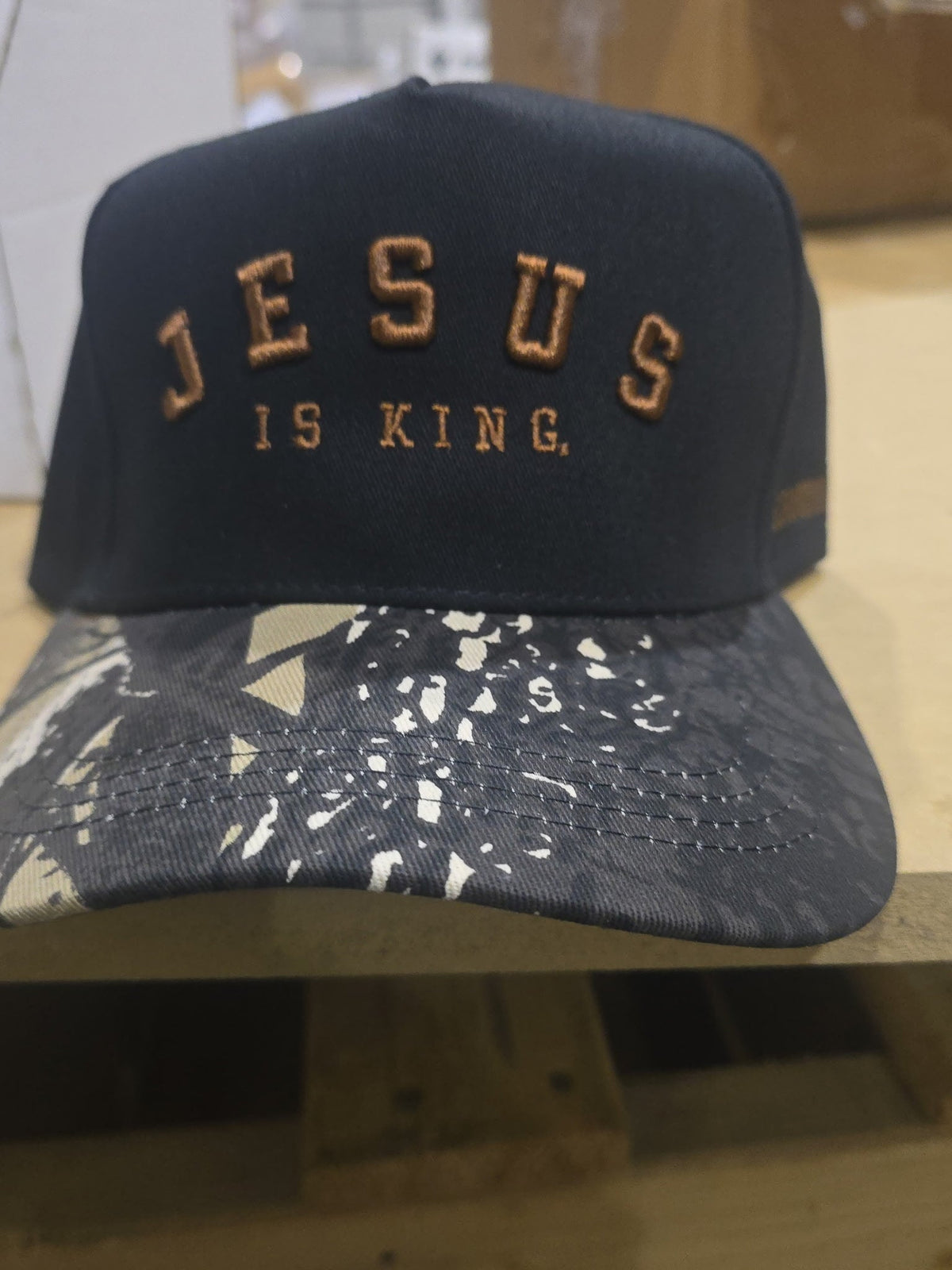 A Born United Private Label Jesus is King - Black and Darkwood Camo Snapback, featuring black fabric embroidered with brown “JESUS IS KING” text and a darkwood camo-patterned brim, rests on a wooden surface.