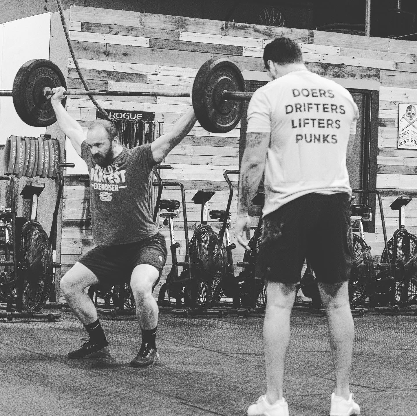 In a black and white gym scene, a man lifts a barbell overhead as another stands before him in the Tango Charlie Apparel Doers Drifters Lifters Punks Unisex Tee—perfect for showing off your crew’s spirit.