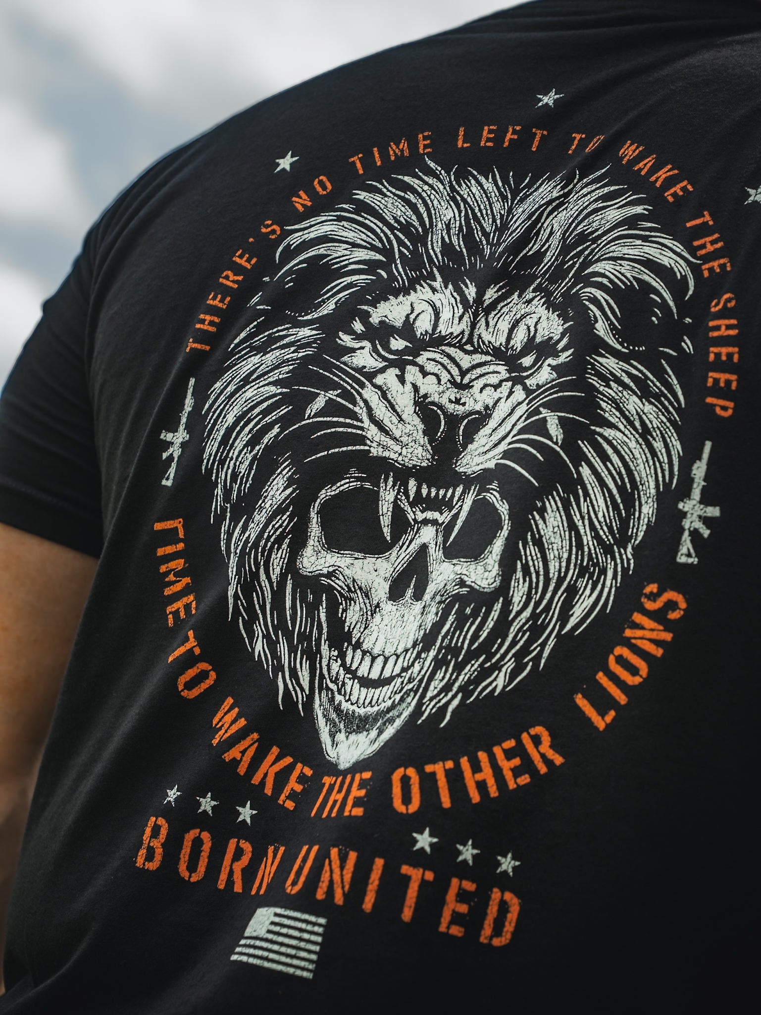A person dons the Waking Lions T-shirt by Born United Private Label, featuring a black tee with a roaring lion graphic above a skull. The text reads, No time left to wake the sheep. Time to wake the other lions, and Born United is displayed above an American flag at the bottom.