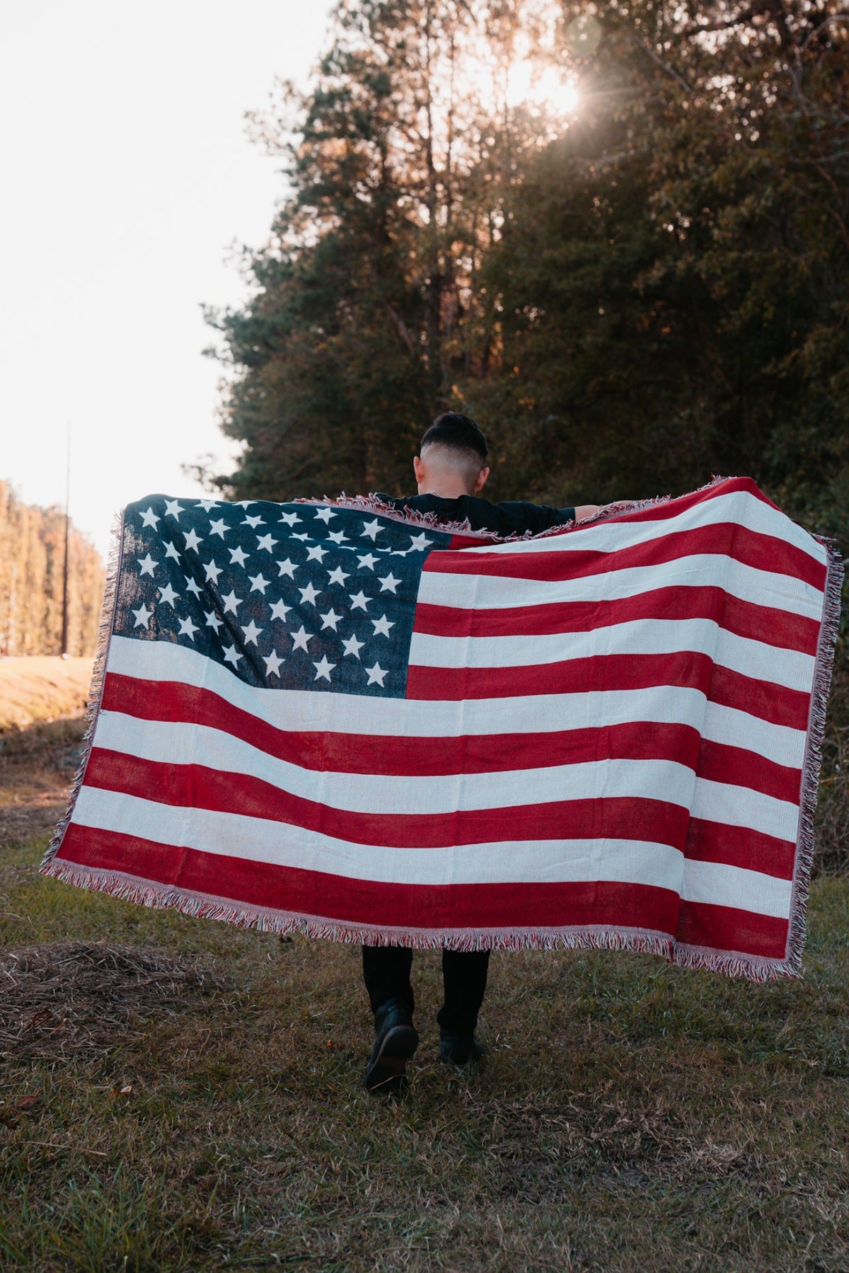 A person walks away on a grassy path, holding an American Flag Jacquard Blanket from Born United Private Label. The sun peeks through tall trees in the background, casting a warm glow on this quintessentially American scene.