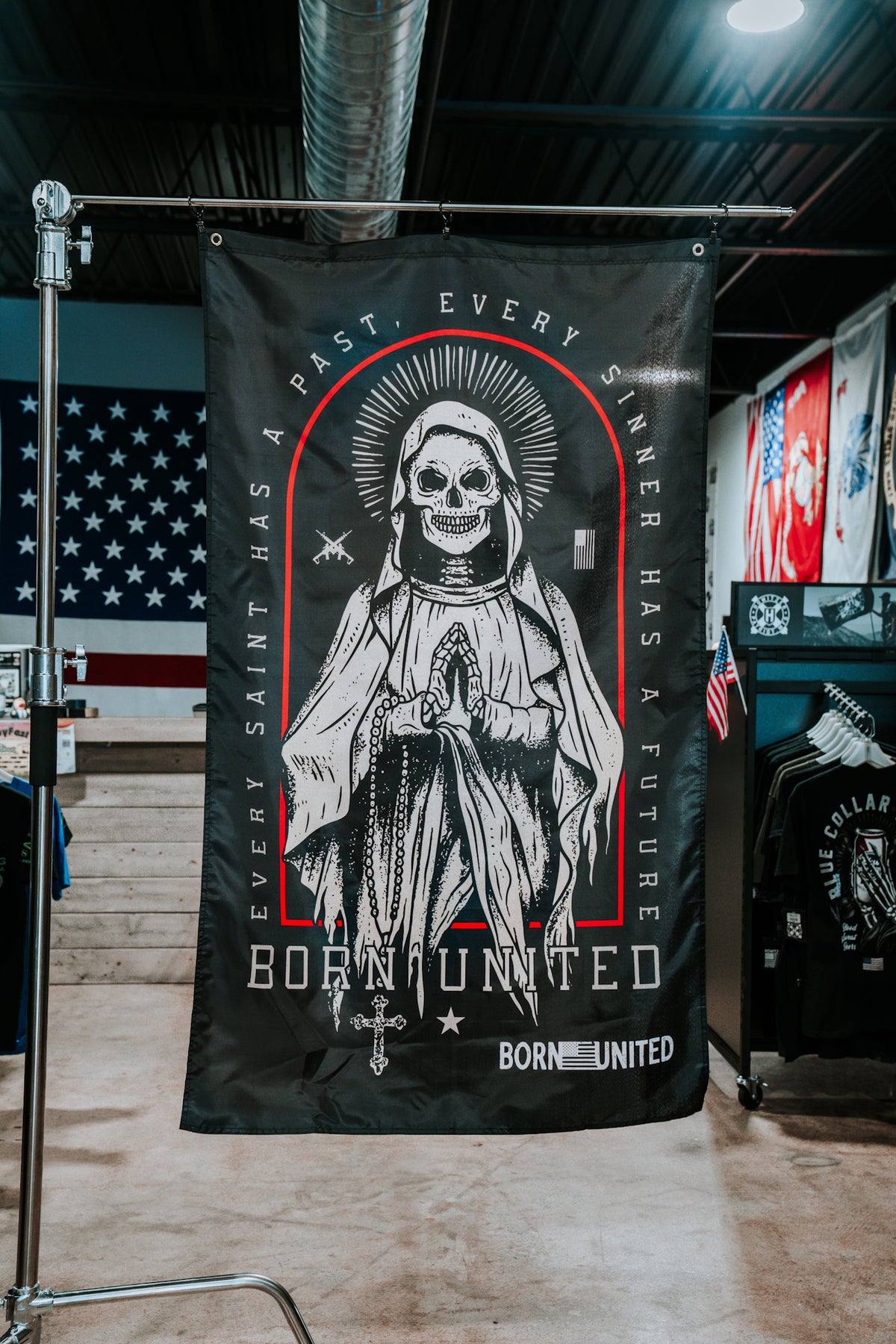 The Saints and Sinners Flag by Born United Private Label is a black polyester tapestry featuring a praying skeletal saint, an American flag background, and the text: Every saint has a past. Every sinner has a future. Born United.