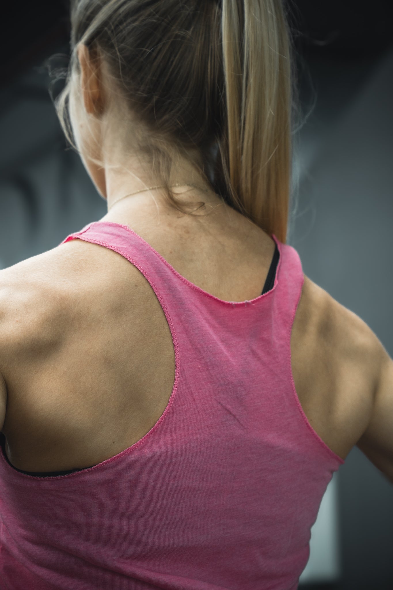 A woman with long blonde hair in a ponytail is seen from behind wearing the Tango Charlie Apparel Lift Heavy. Pet Dogs. womens pink tank, showing off her back and shoulder muscles against a blurred dark background.