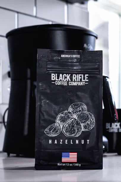 A black bag of Black Rifle Coffee Hazelnut Coffee Roast - Ground sits on a kitchen counter, exuding bold flavor. The packaging displays hazelnut illustrations and an American flag, with a coffee maker blurred in the background.