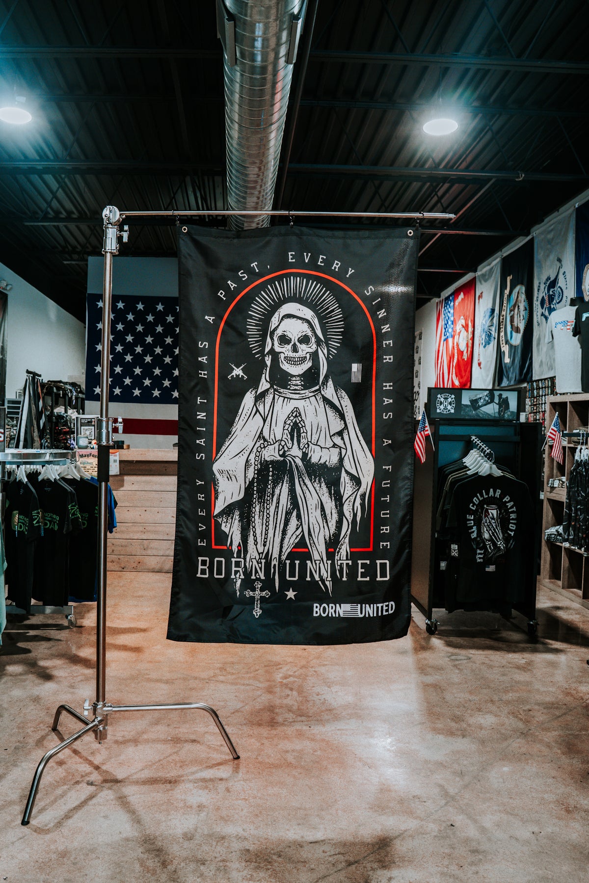 The Saints and Sinners Flag by Born United Private Label is a large black polyester banner featuring a skeleton saint with halo and praying hands, displaying the text: EVERY SAINT HAS A PAST, EVERY SINNER HAS A FUTURE. Various clothes and flags are nearby.