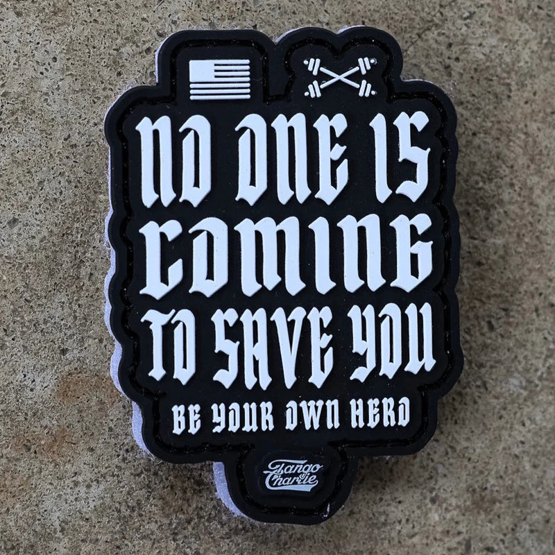 The No One Is Coming To Save You PVC Patch by Tango Charlie Apparel features white gothic text, a small flag, and crossed barbells above the message. The patch displays the Tango Charlie logo at the bottom and is shown on a stone surface.