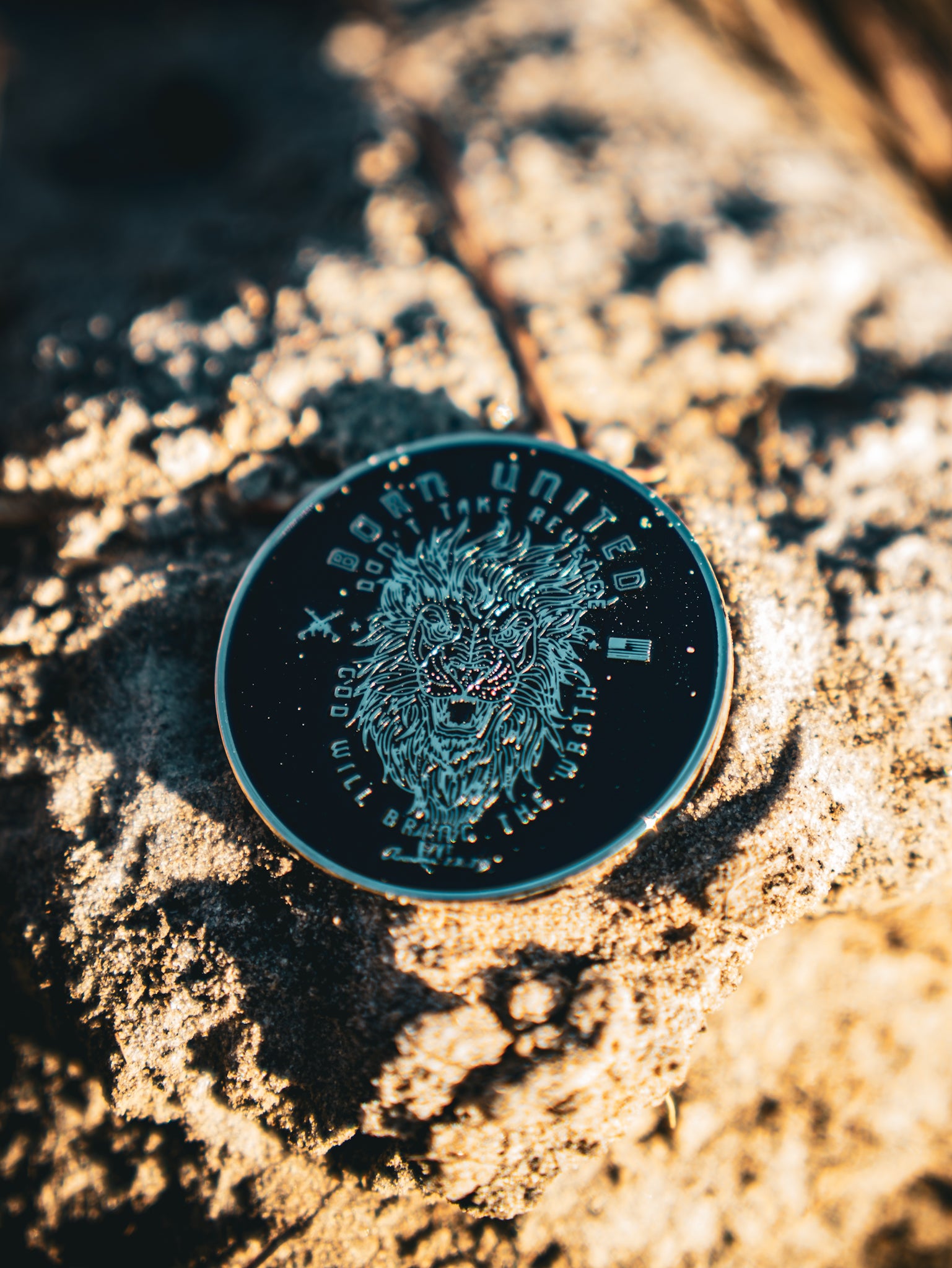 The Revenge Challenge Coin by Born United Private Label, featuring a roaring lion, rests on a textured stone surface. Its intricate engravings are beautifully illuminated by sunlight, creating striking shadows.