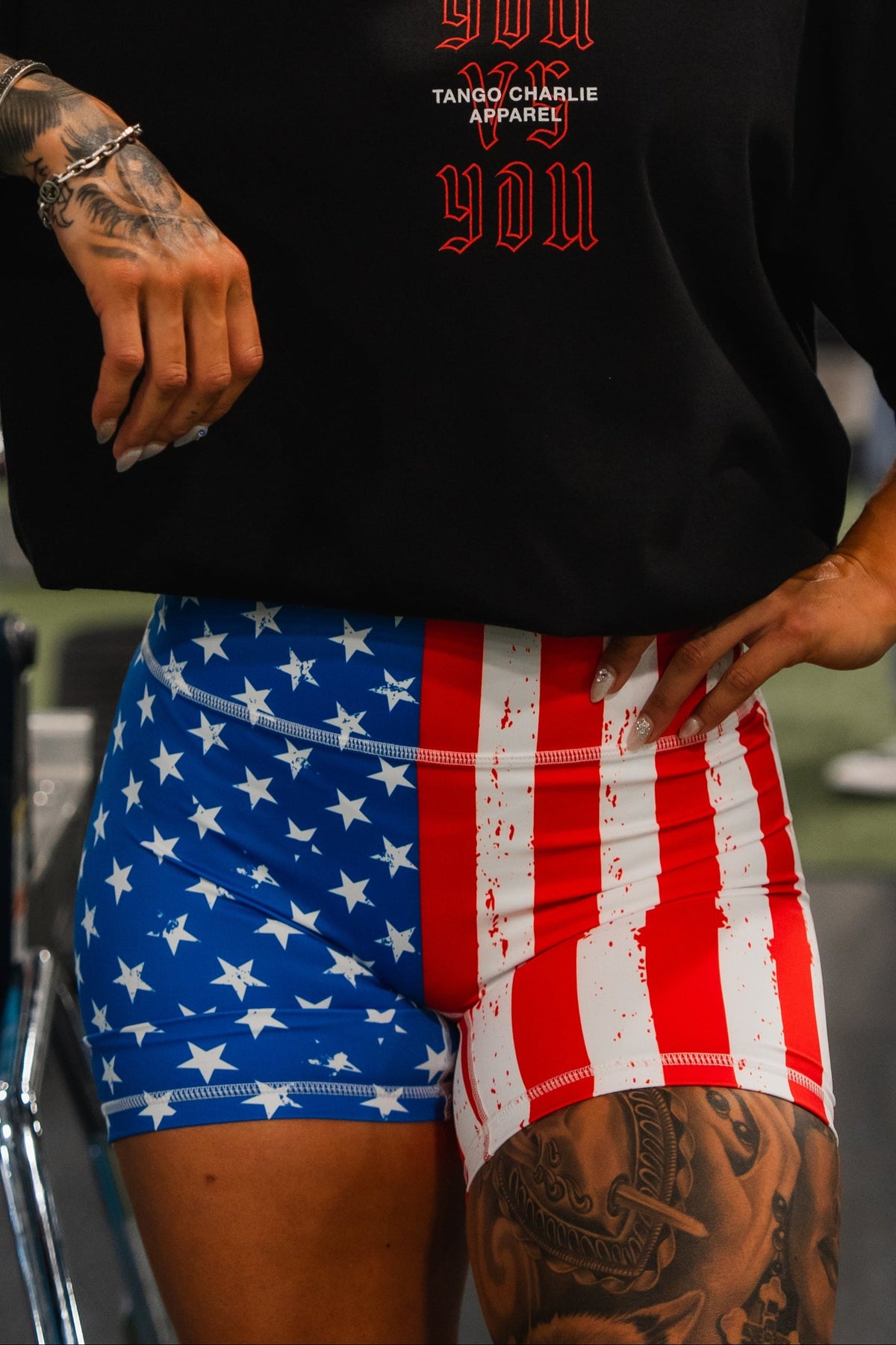 A tattooed woman stands in a gym, resting her arm on blue equipment. She wears a black T-shirt and Born United Private Label Women’s High Waisted Seamless Front Compression Training Shorts | Stars & Stripes. Other people and gym gear are visible.