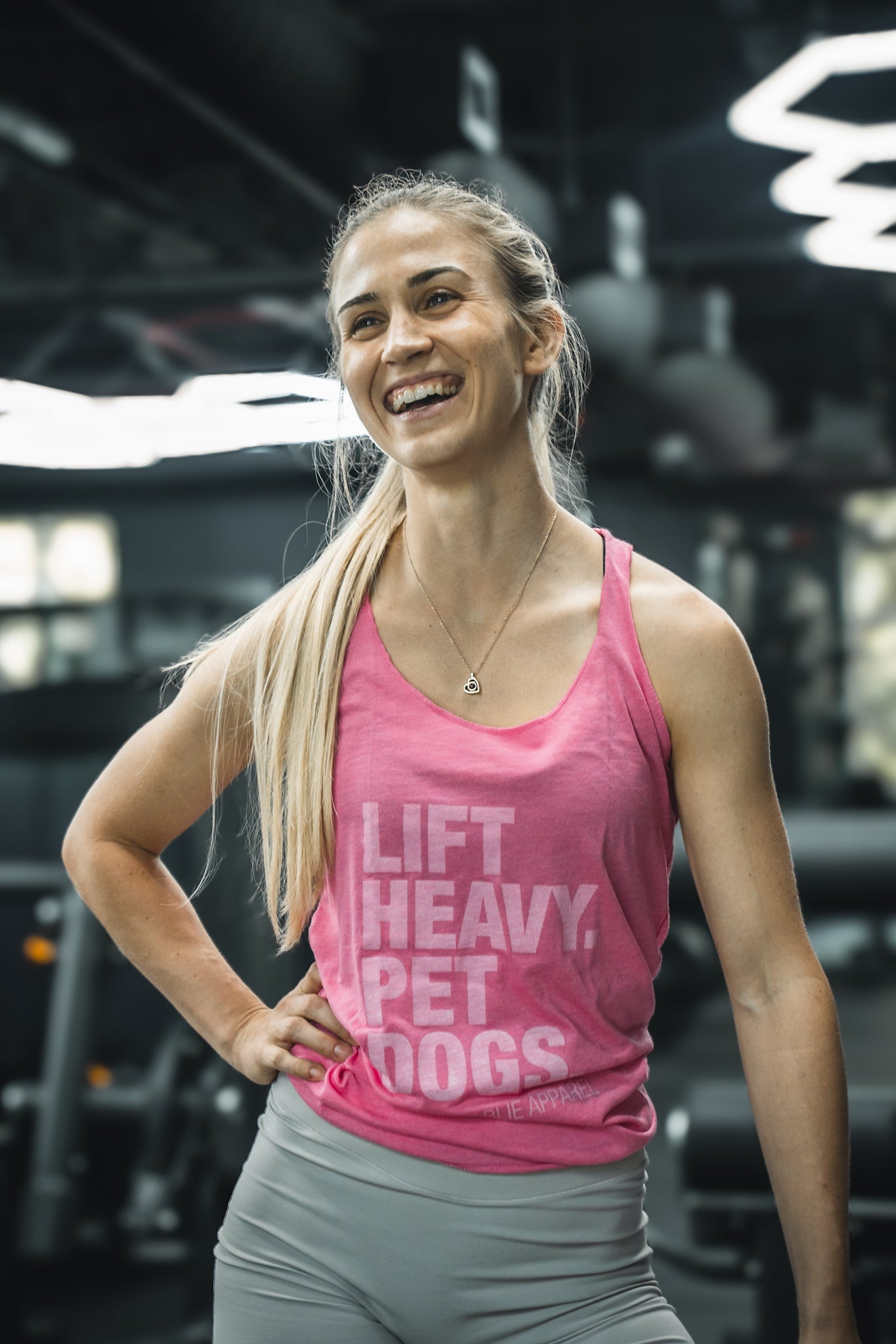 A smiling woman with long blonde hair in a ponytail stands in a gym, wearing the Tango Charlie Apparel Lift Heavy. Pet Dogs. Womens Pink Tank and light gray leggings, with gym equipment visible in the background.