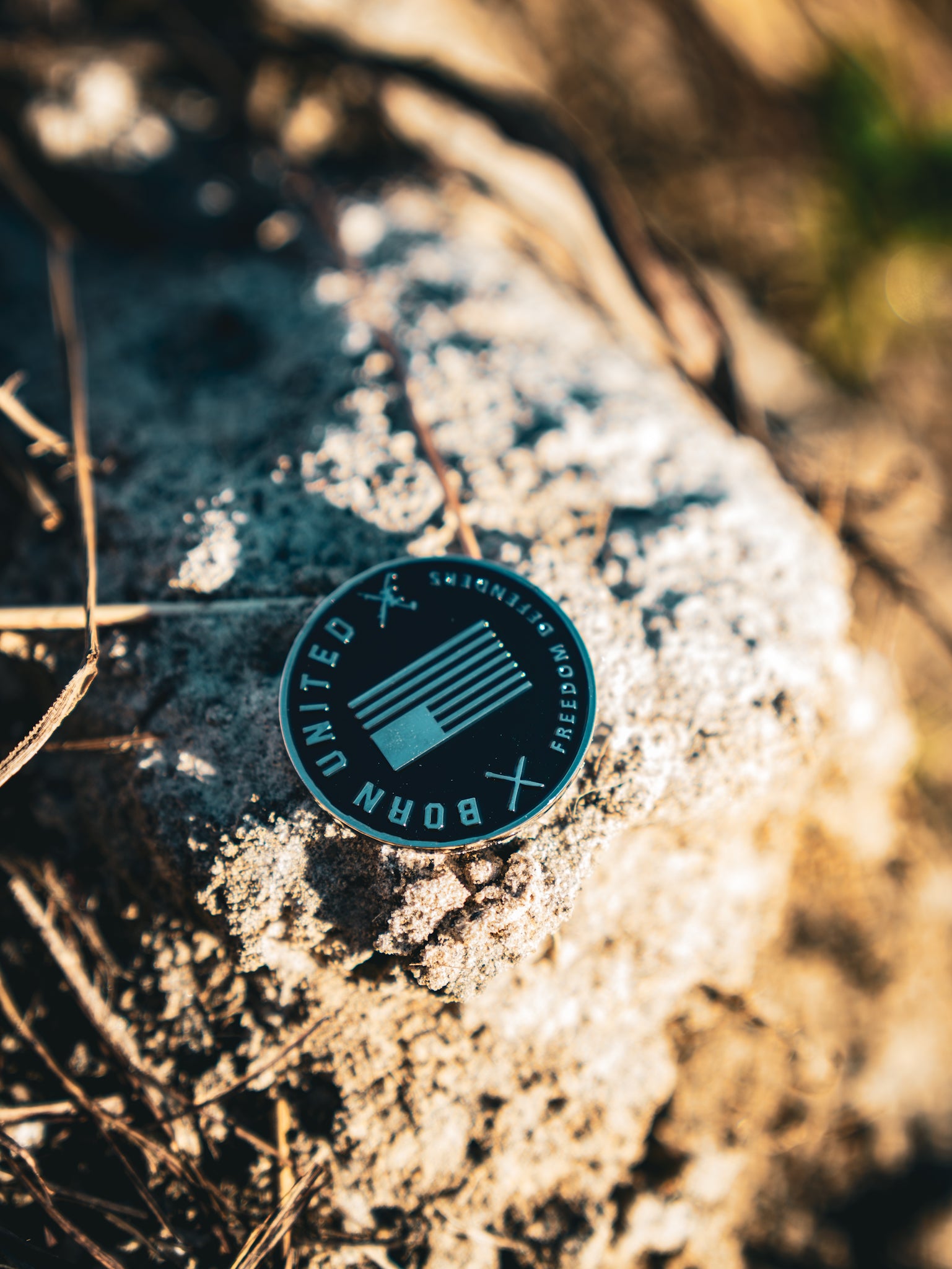 The Revenge Challenge Coin by Born United Private Label, resembling a round black badge with an abstract design and BORN UNITED, sits on textured rock. Sunlight and shadows from nearby dried grass enhance this rustic, natural setting.
