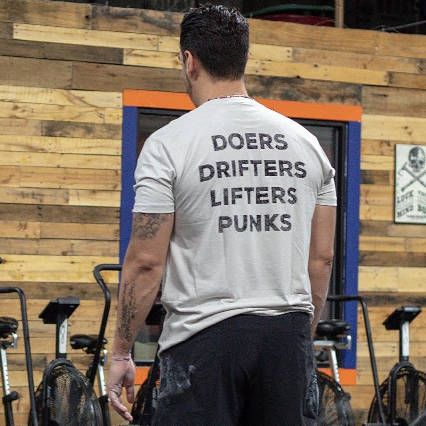 In a gym, a man wears the Tango Charlie Apparel Doers Drifters Lifters Punks Unisex Tee—white with bold back print. Exercise bikes and a wood wall are behind him. Show off your crew with this standout fitness shirt.