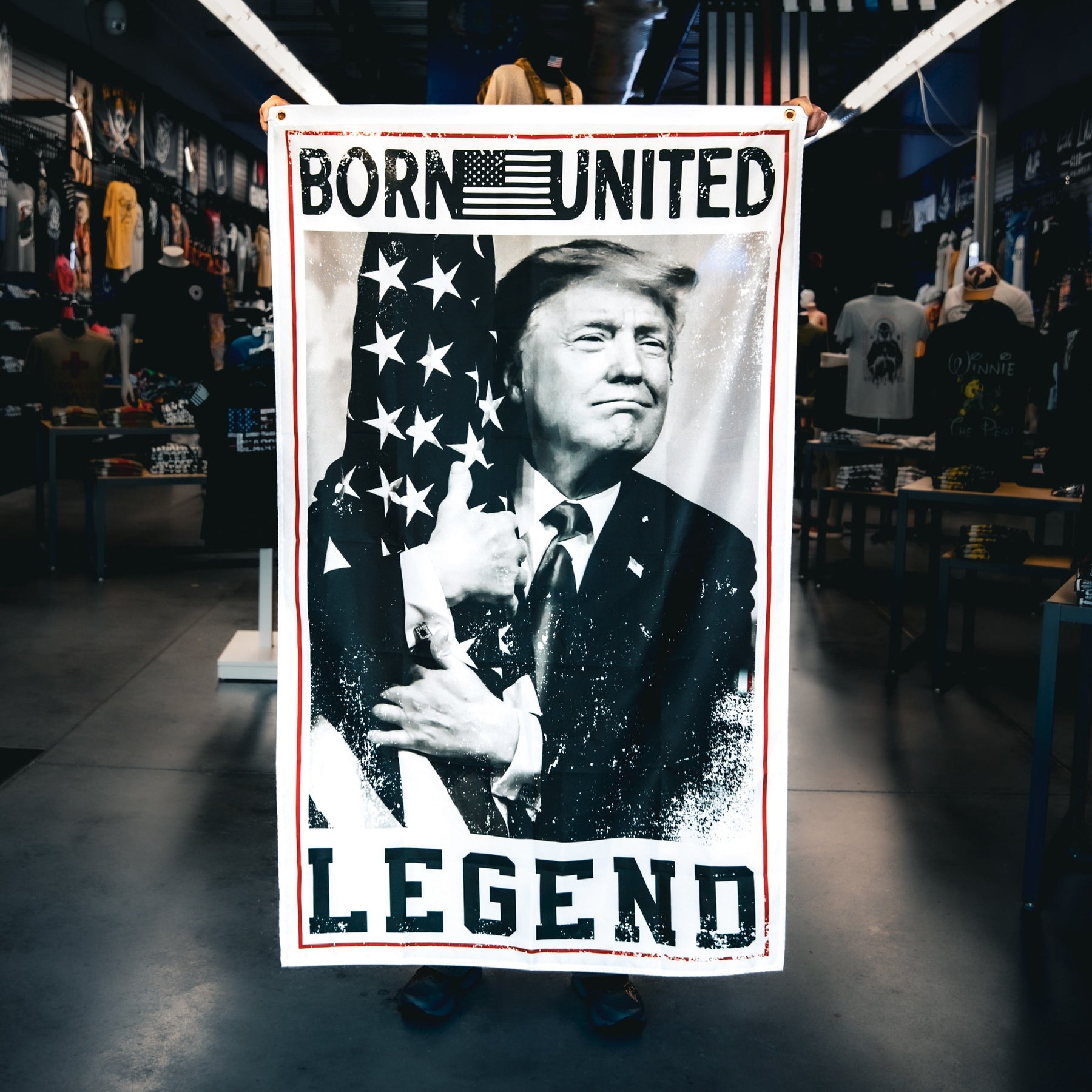 A person holds up a blanket featuring a black-and-white image of a man hugging the American flag, with the words BORN UNITED LEGEND at the top and bottom. The setting is a store with various merchandise on display.