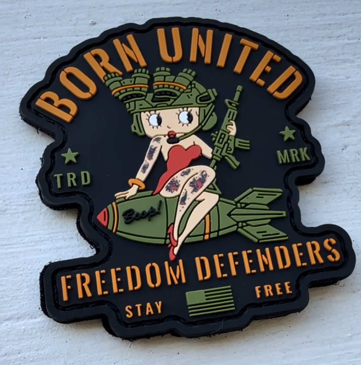 The Betty Boop PVC Patch by Born United Private Label features a cartoon pin-up in military gear, riding a missile. It embodies Freedom Defenders with the text Born United, TRD MRK, and Stay Free as Betty Boop wears a red outfit with a military-style helmet.