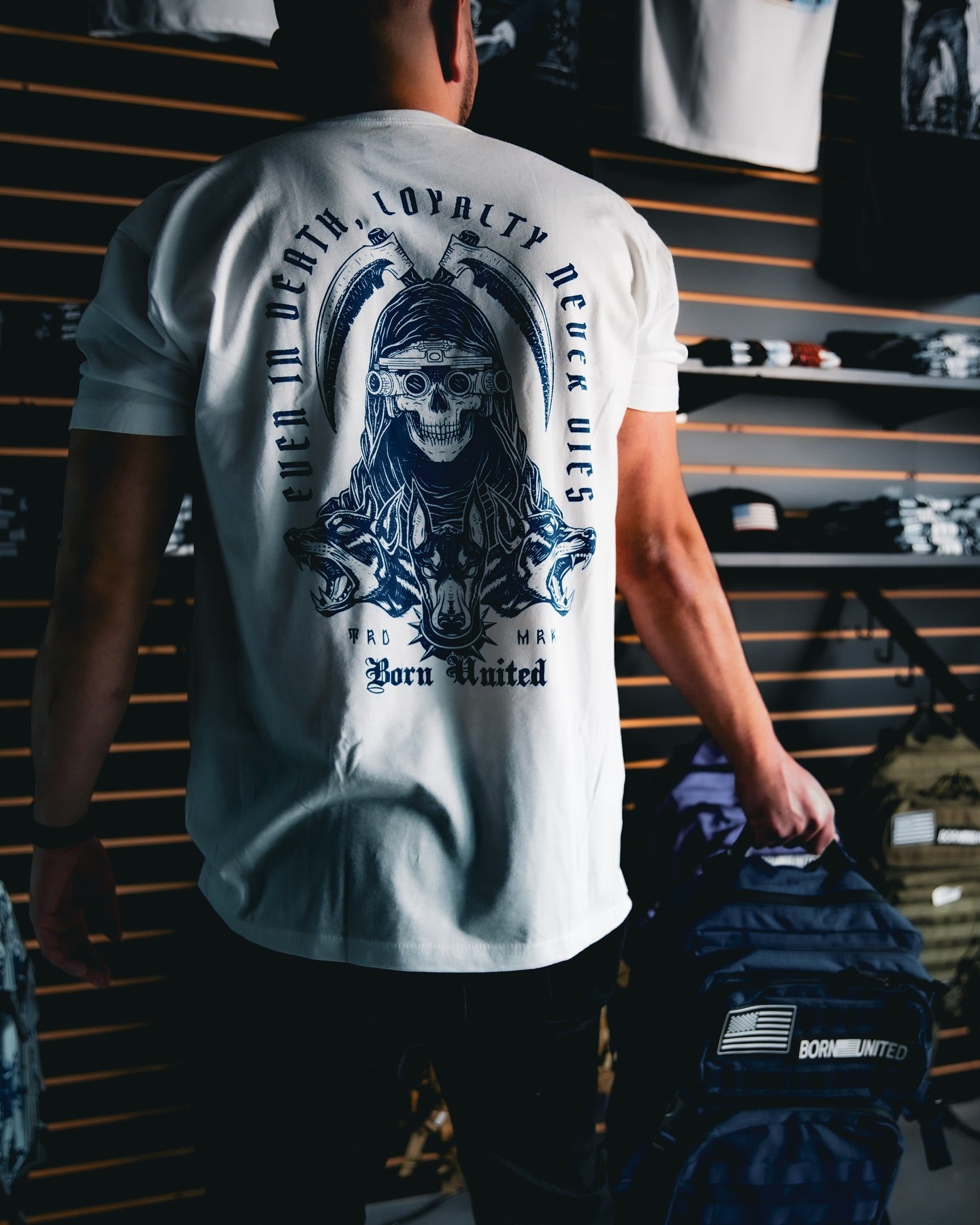 Man wearing a white t-shirt with a large black graphic on the back featuring a skull design. The shirt has the words Loyalty Never Dies. He is holding a blue backpack with an American flag patch. The background shows clothes on shelves.