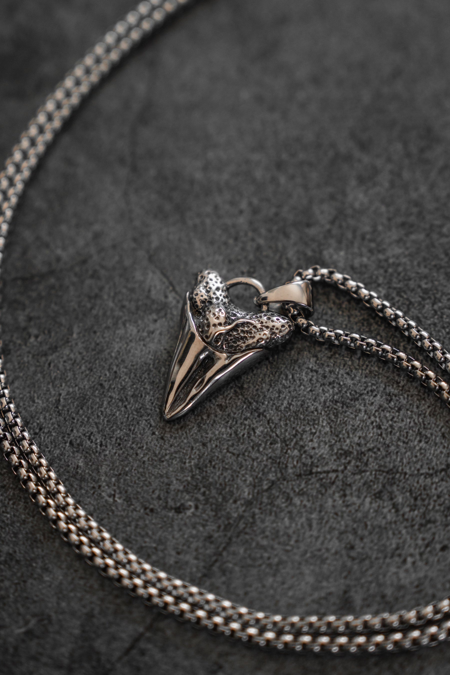The Relentless Predator Necklace by Relentless Betrayal features a silver heart-shaped pendant with intricate detailing, hanging on a textured stainless steel chain set against a dark gray background.