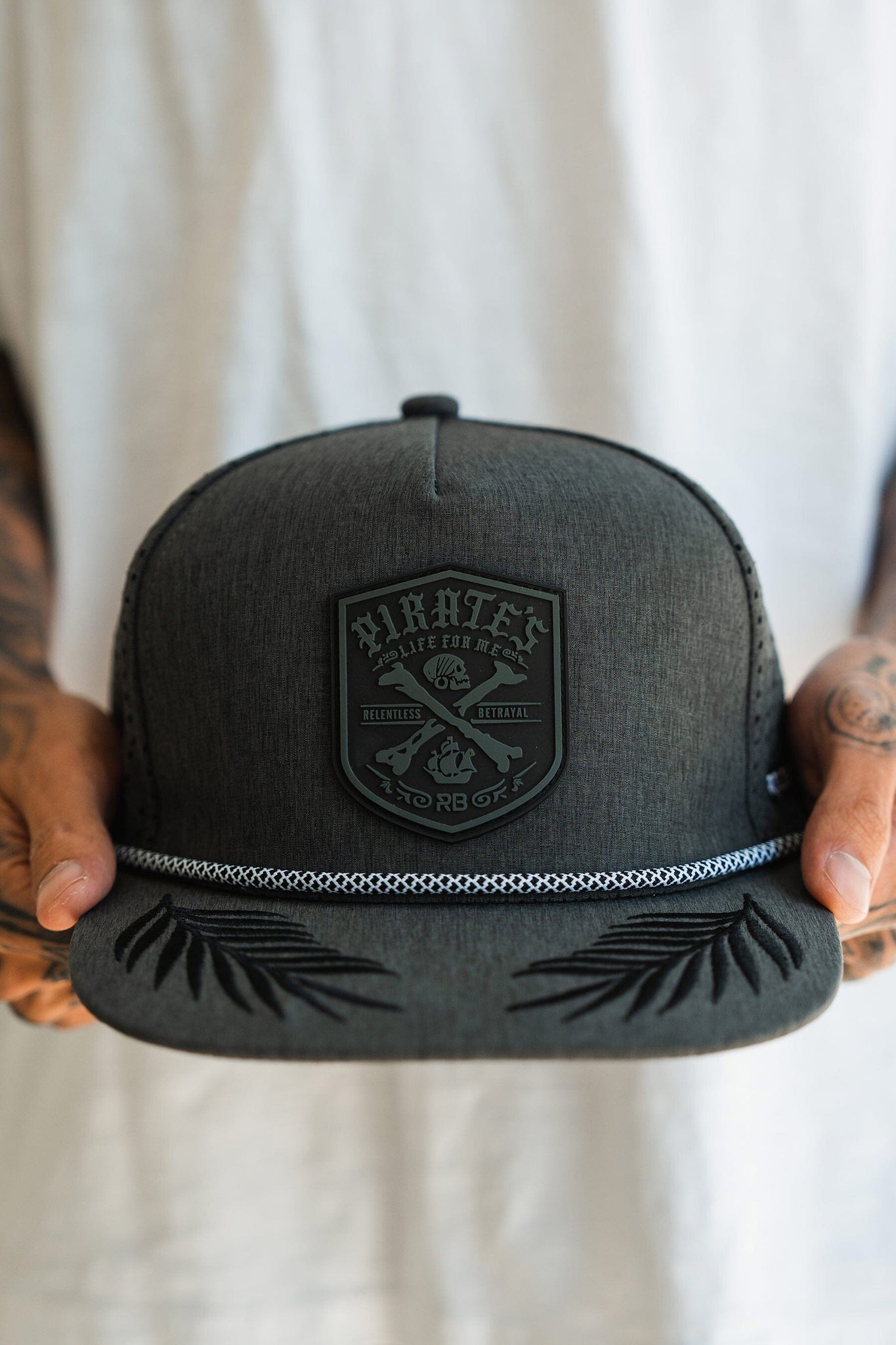 A person holds the Pirates Life Grey Snapback from Relentless Betrayal, featuring a dark gray design with a PVC rubber patch showing crossed swords. The detailed visor adds character, and a white shirt is visible in the background.