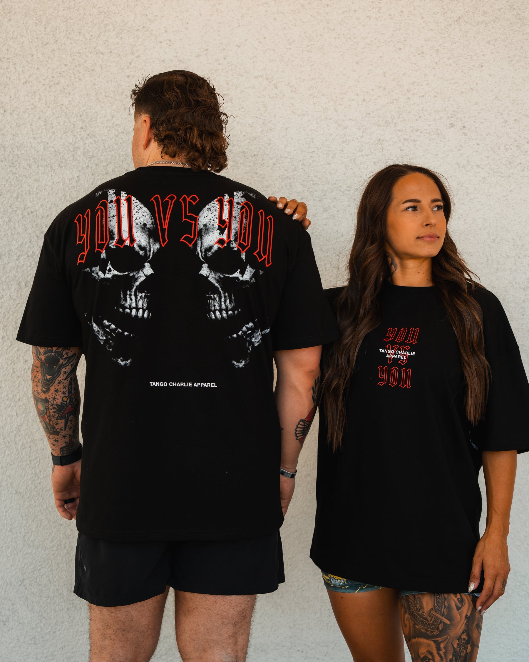 A man and woman wear Tango Charlie Apparel’s You vs You - Premium Oversized Tee. The man displays the skull back graphic, while the woman faces forward, showing the unisex tee’s front design against a plain wall.