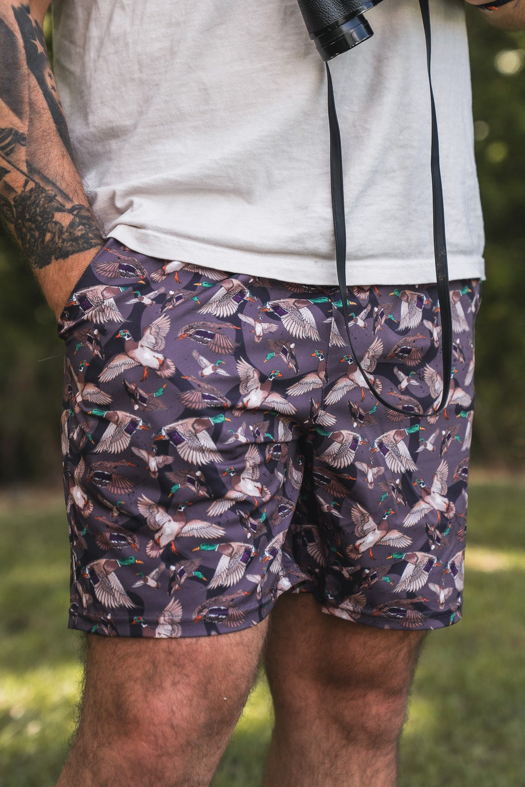 A tattooed individual stands outdoors in a white t-shirt and Mucker Bucket Men's Performance Athletic Shorts | 5.5" – Duck Wild, featuring a colorful bird pattern, with binoculars in hand amid a serene, green backdrop.