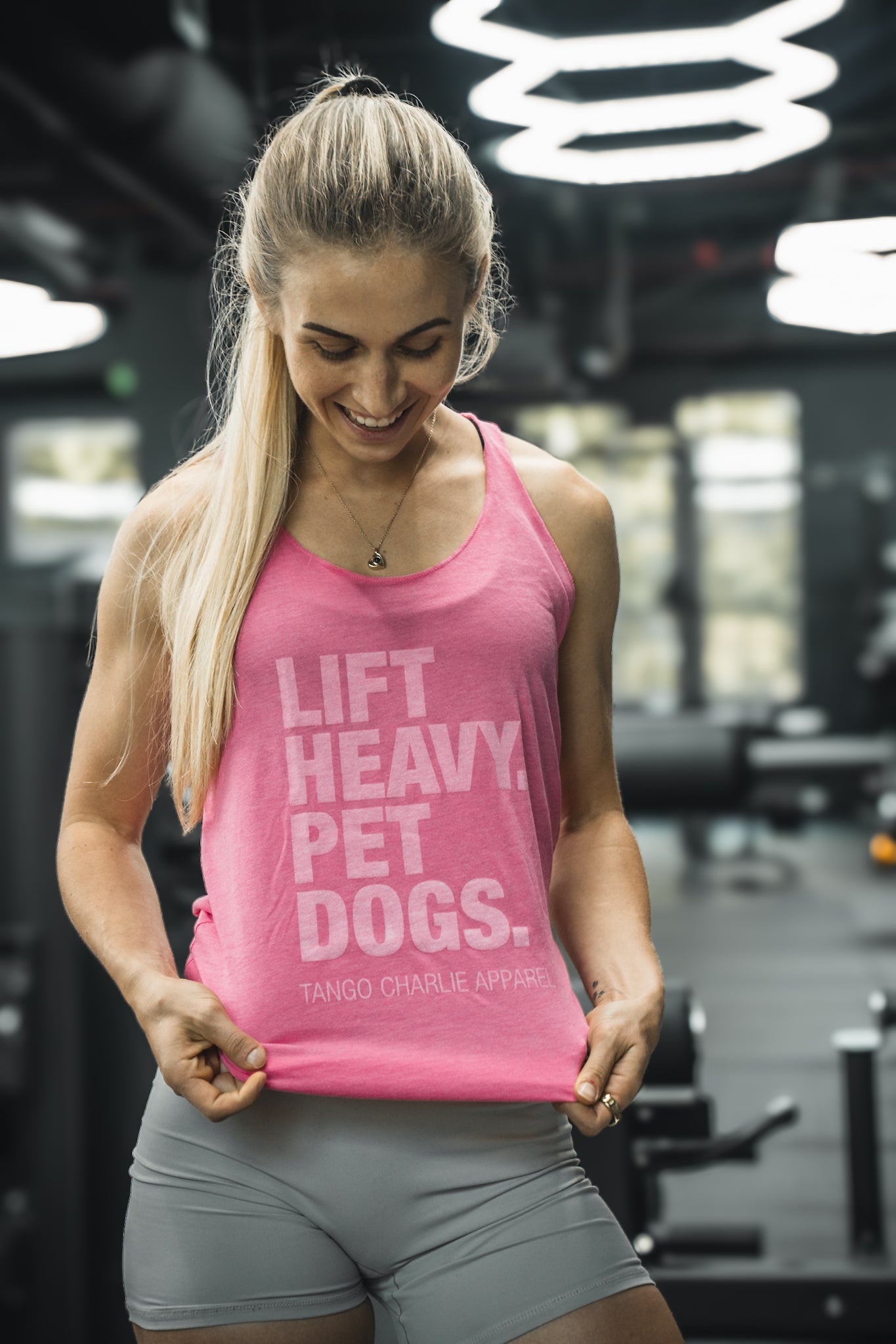 A woman at the gym smiles, holding her pink Lift Heavy. Pet Dogs. womens tank by Tango Charlie Apparel, featuring vintage-style ink. She pairs it with gray shorts and stands in a modern fitness facility.