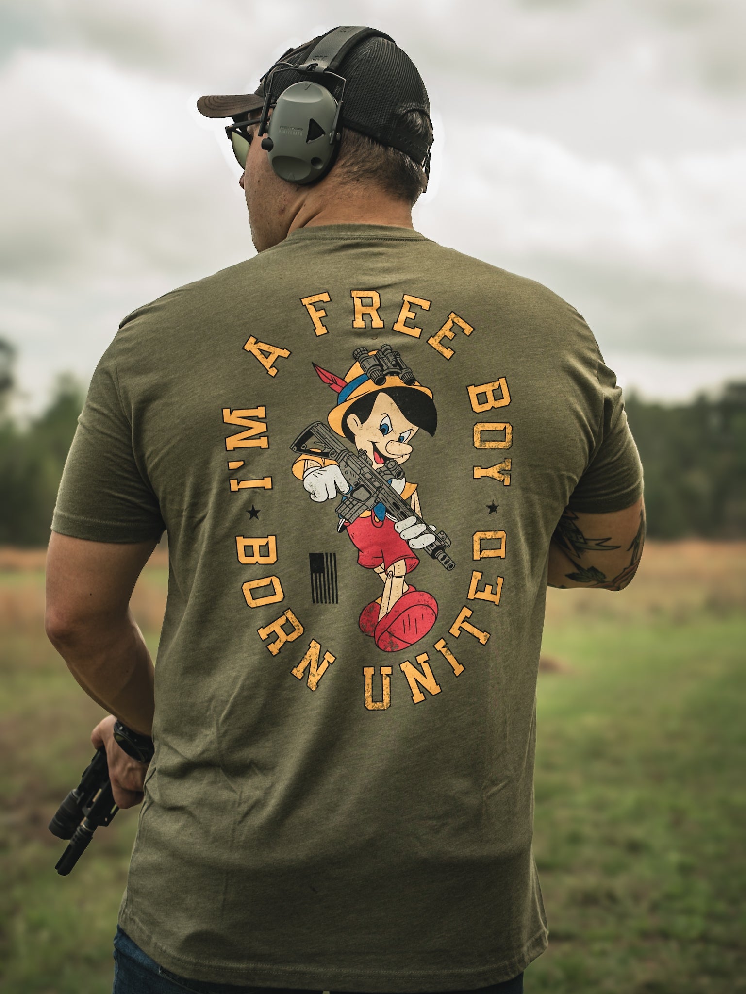 A man outdoors sports headphones and an olive green Free Boy Tee by Born United Private Label, featuring a cartoon character with a gun. The surrounding text reads, Im a free boy but united. Standing in the grass, he confidently holds a gun.
