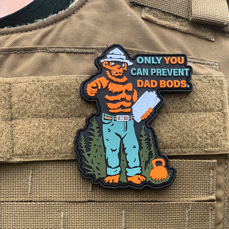 The Dad Bods Patch from Tango Charlie Apparel features a cartoon bear in jeans and a hat, lifting a dumbbell, with the message ONLY YOU CAN PREVENT DAD BODS. It’s designed with velcro for easy attachment to any tactical vest.