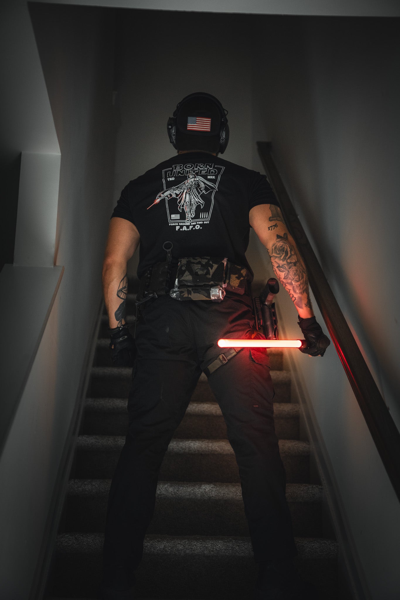 A person in tactical gear sports the Born United Private Label Vader FAFO tee, with tattooed arms, a helmet featuring an American flag patch, and a glowing red baton while standing on a staircase.