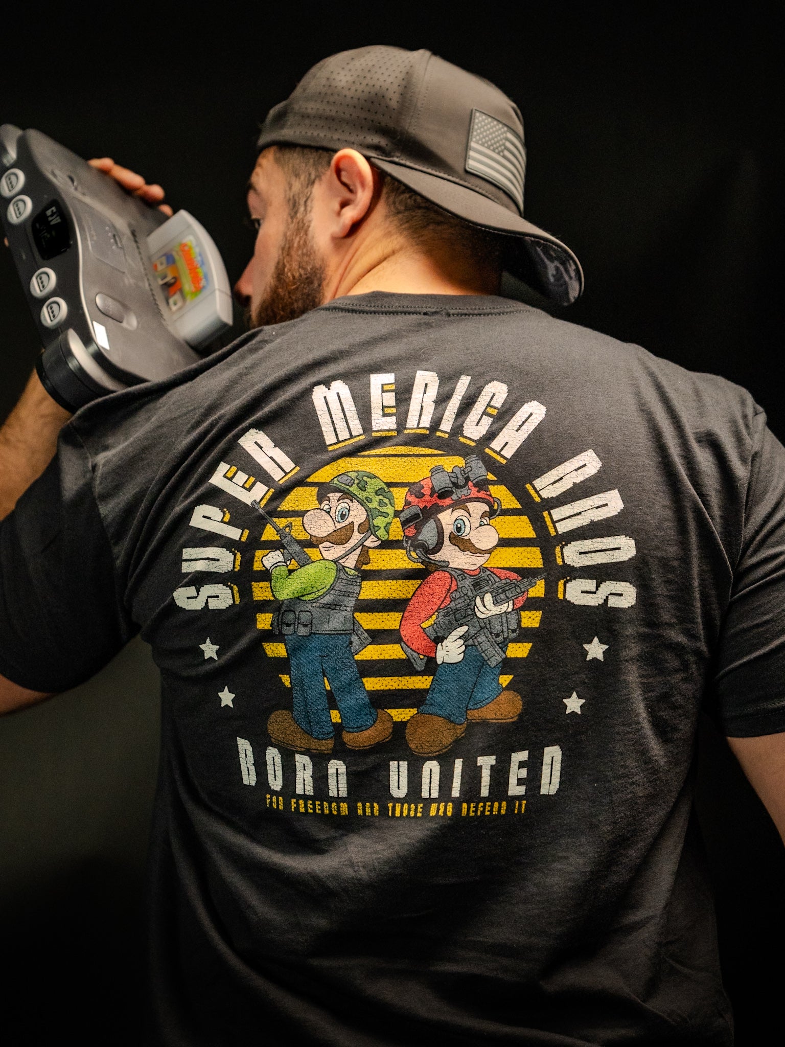 A man in a backwards cap holds an old video game console to his mouth. Hes wearing a premium tee from Born United Private Label, called Super Merica Bros, featuring cartoon plumbers and the slogan For Freedom and Those Who Defend It, celebrating an all-American update of Brothers In Arms.
