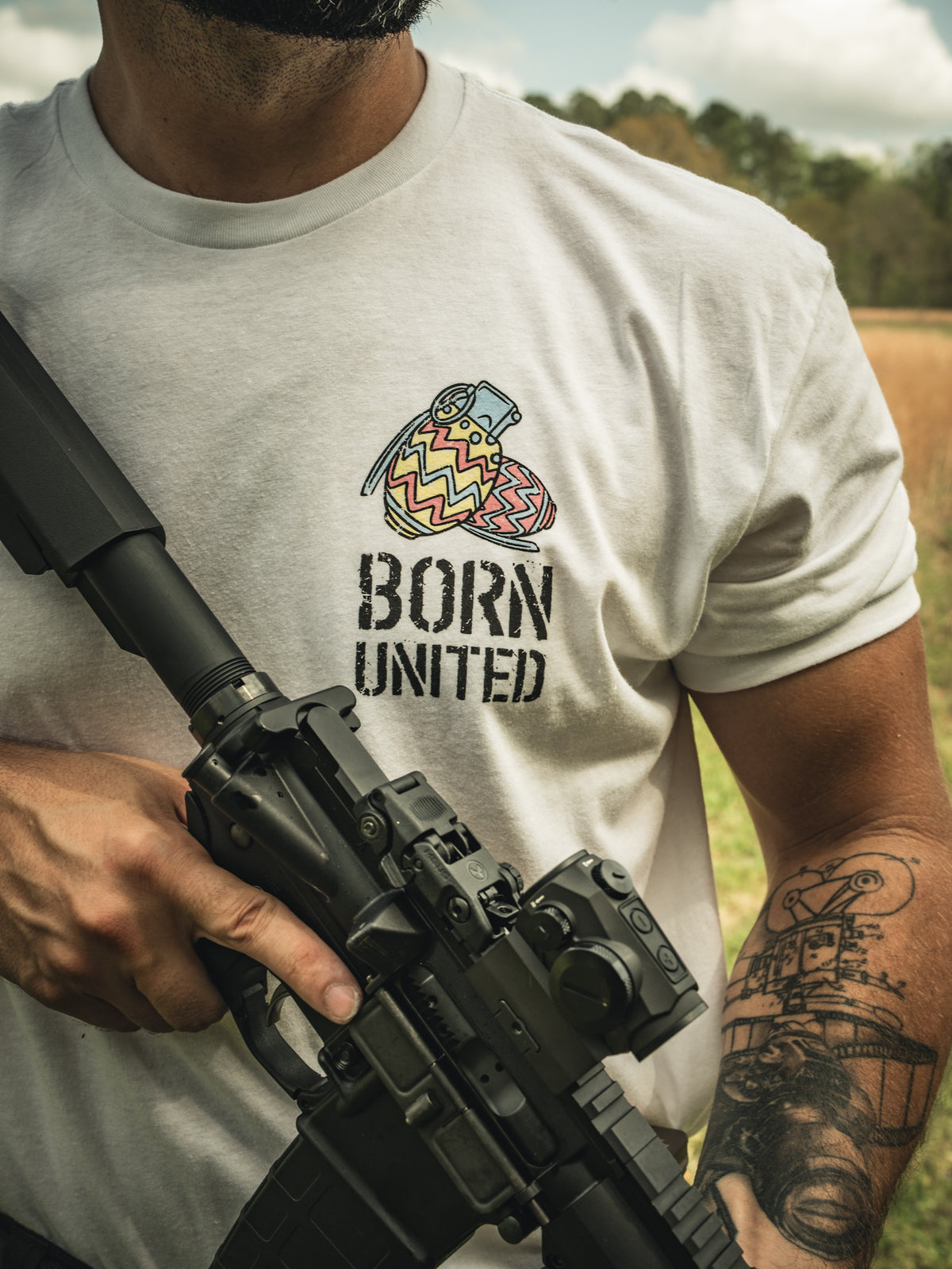 Outdoors, a person holds a rifle and wears a white Born United Private Label "Battle Bunny" t-shirt with heart and bandage art. They have a forearm tattoo, standing on grass with trees and a partly cloudy sky in the background.