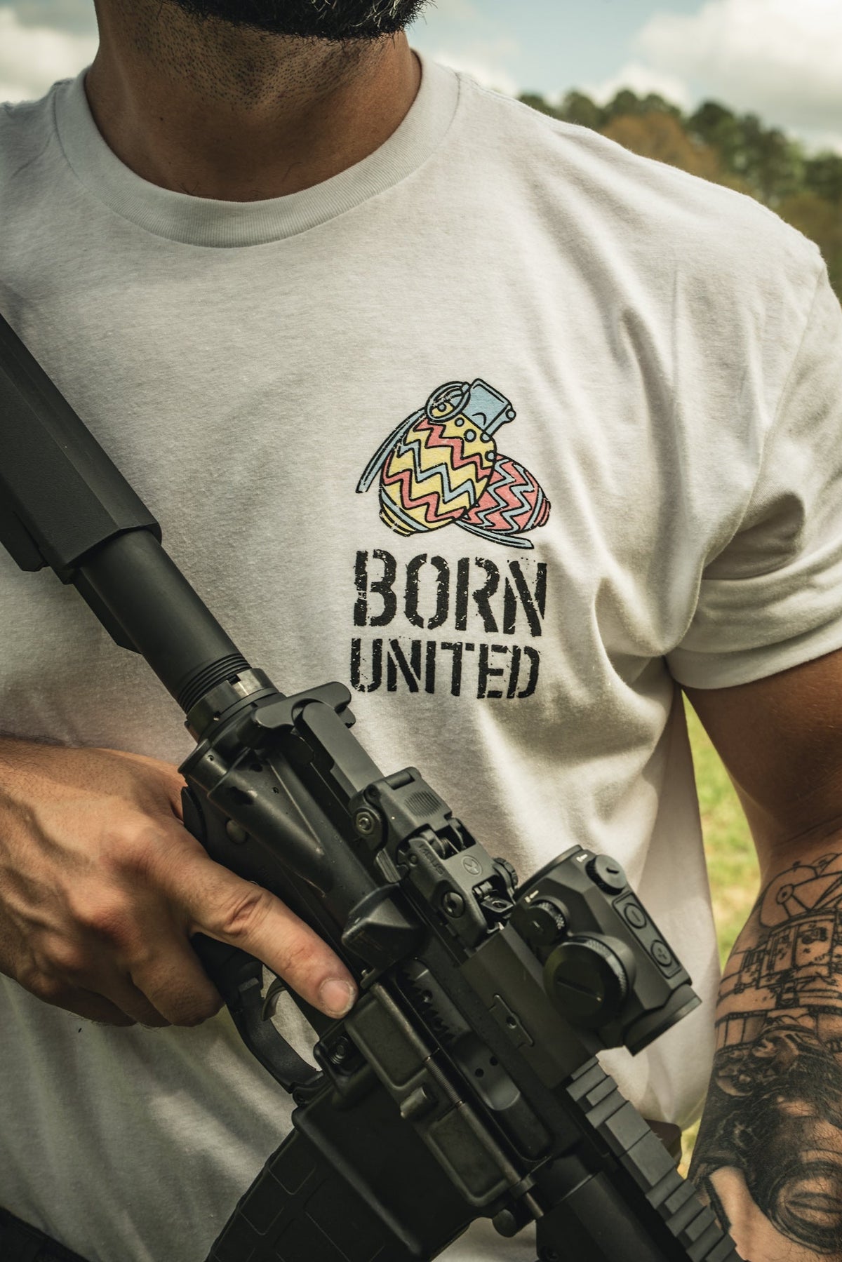 Outdoors, a person holds a rifle and wears a white Born United Private Label "Battle Bunny" t-shirt with heart and bandage art. They have a forearm tattoo, standing on grass with trees and a partly cloudy sky in the background.