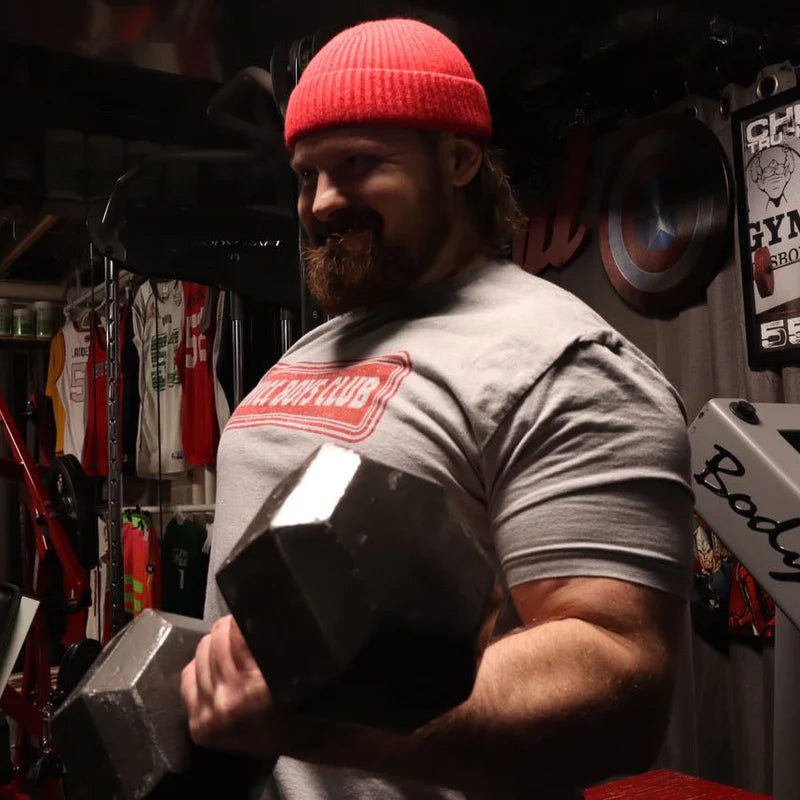 A bearded man in a red beanie lifts a large dumbbell in the gym, smiling. He wears a gray THICC Boys Club T-Shirt by Tango Charlie Apparel, with weights and plates energizing the background.