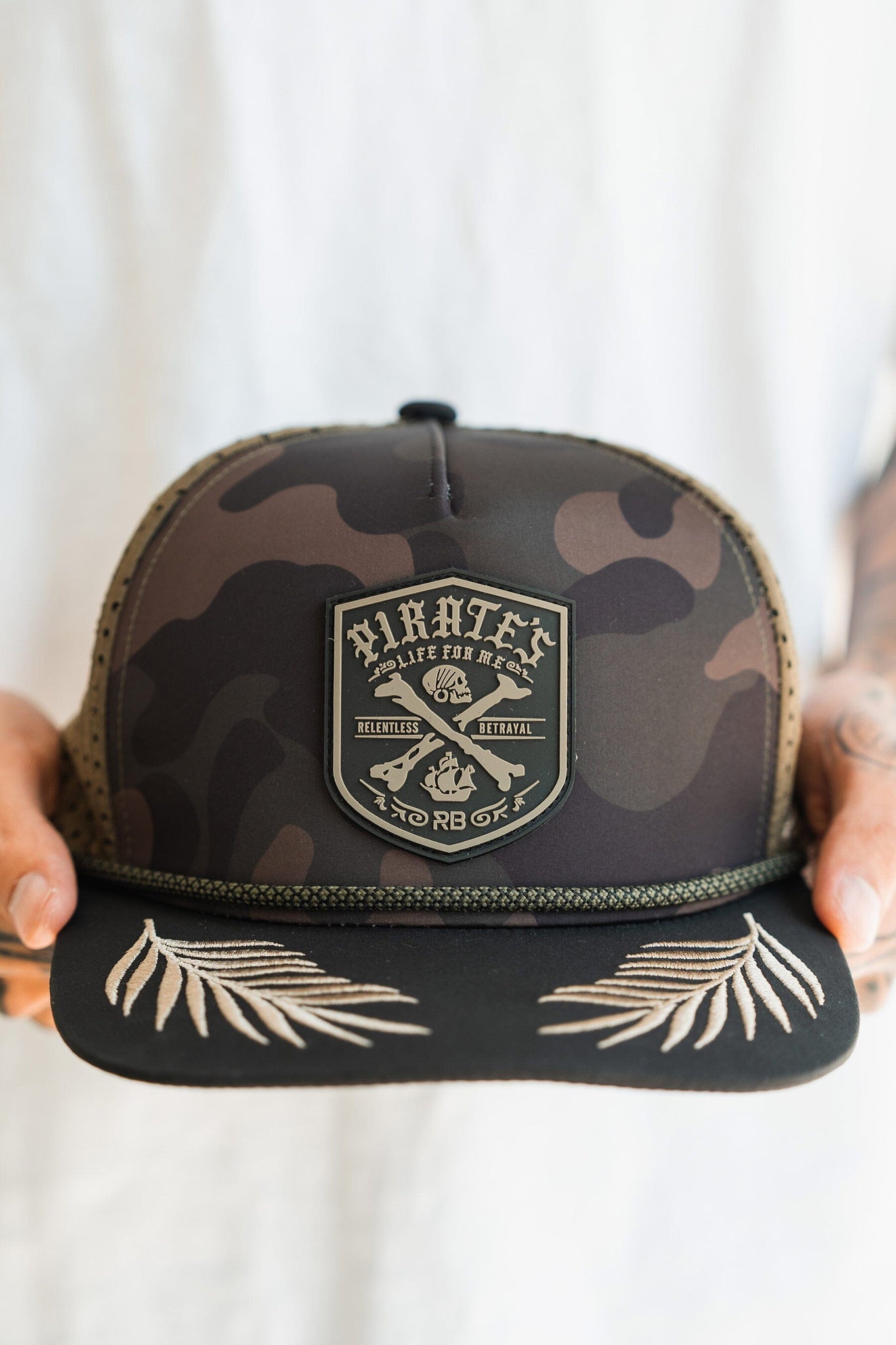 A person holds the Pirates Life Camo Snapback by Relentless Betrayal, showcasing a detailed Pirates, Live for the Hunt, No Rb Off patch with crossed rifles and skull. Its brim is embroidered with palm leaf designs—ideal for adventurers embracing a pirates life.